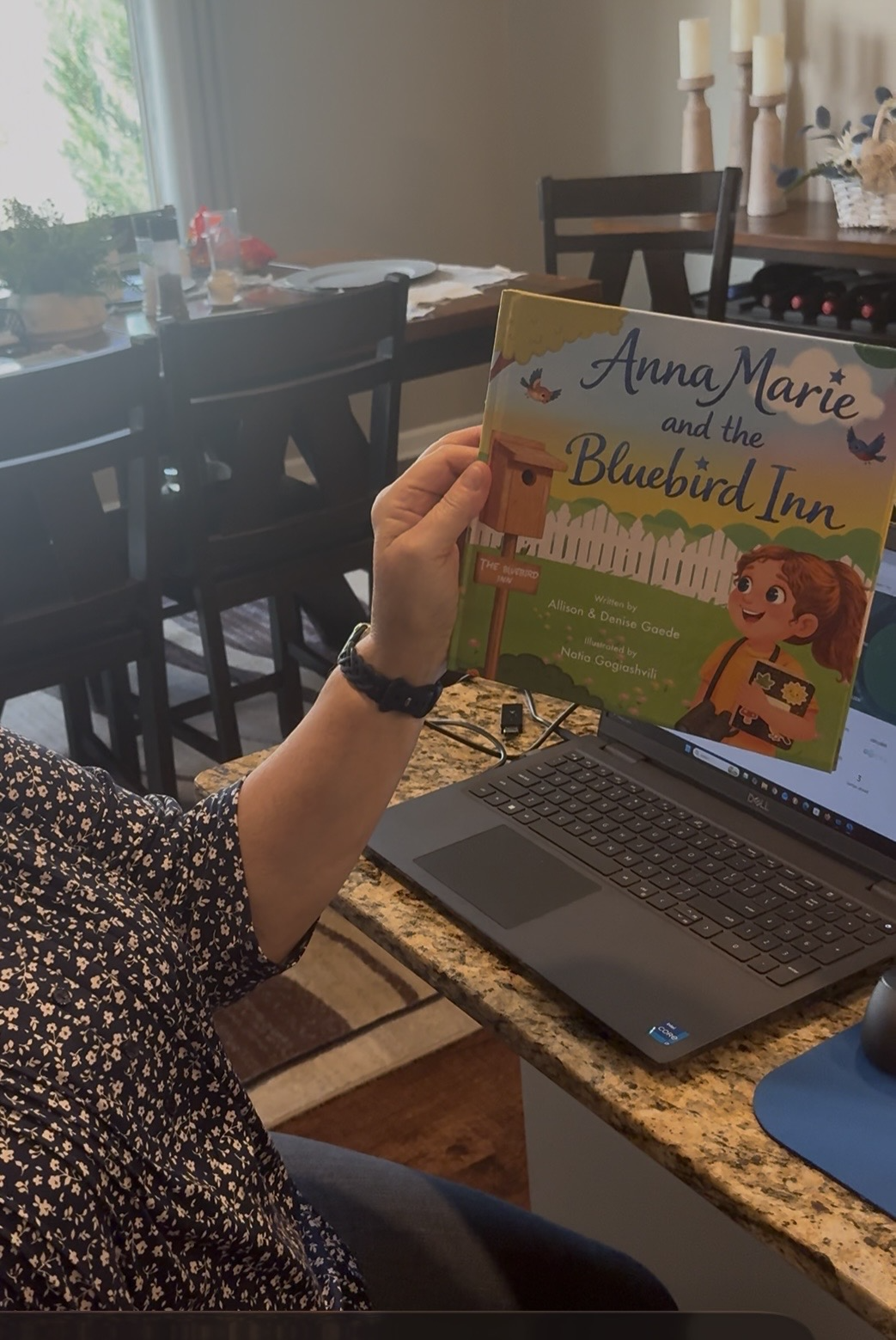 Person sitting at a kitchen counter holding a children's book titled 'Anna Marie and the Bluebird Inn' with a house and garden illustration, a laptop on the counter, and a dining table set in the background.