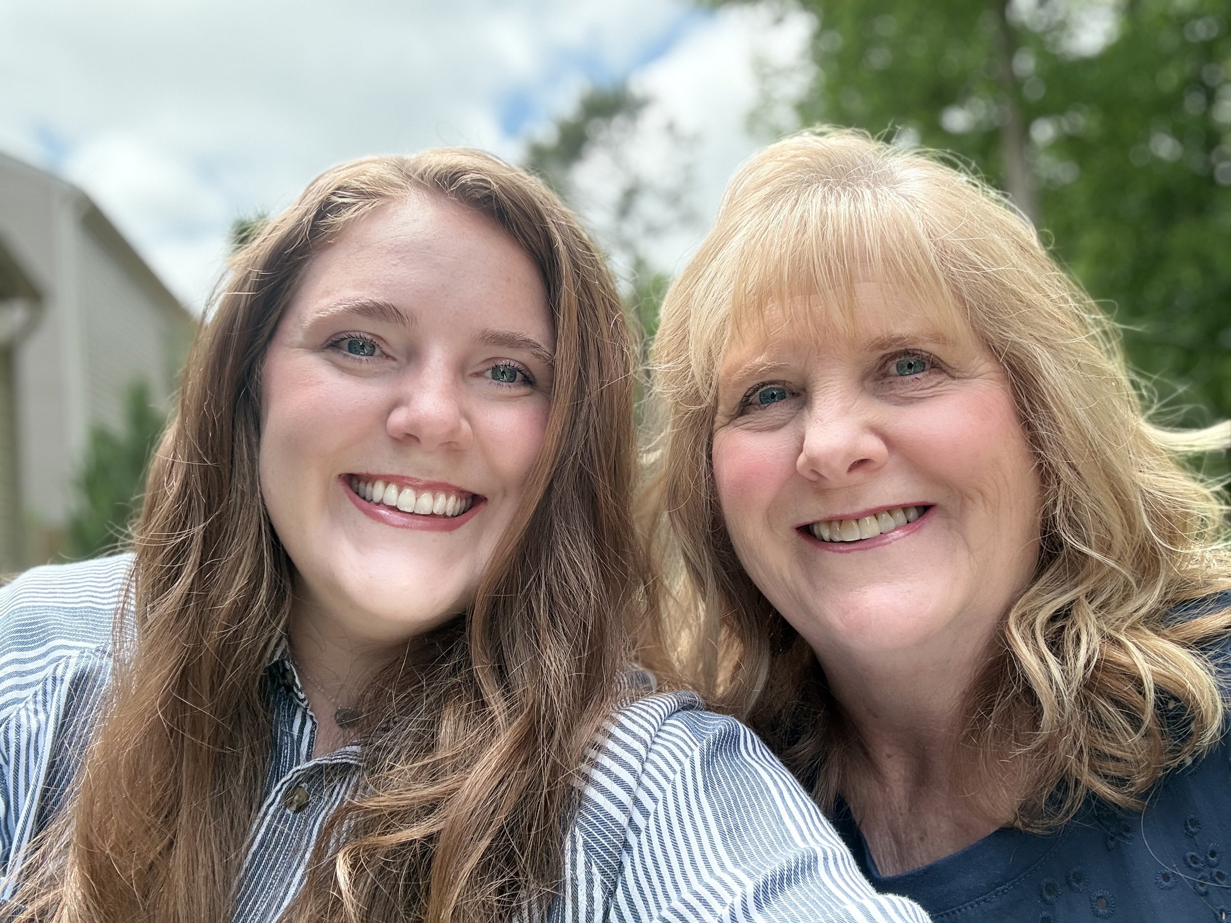 Close-up of two smiling women with long hair, outdoors with cloudy sky and green trees in the background.