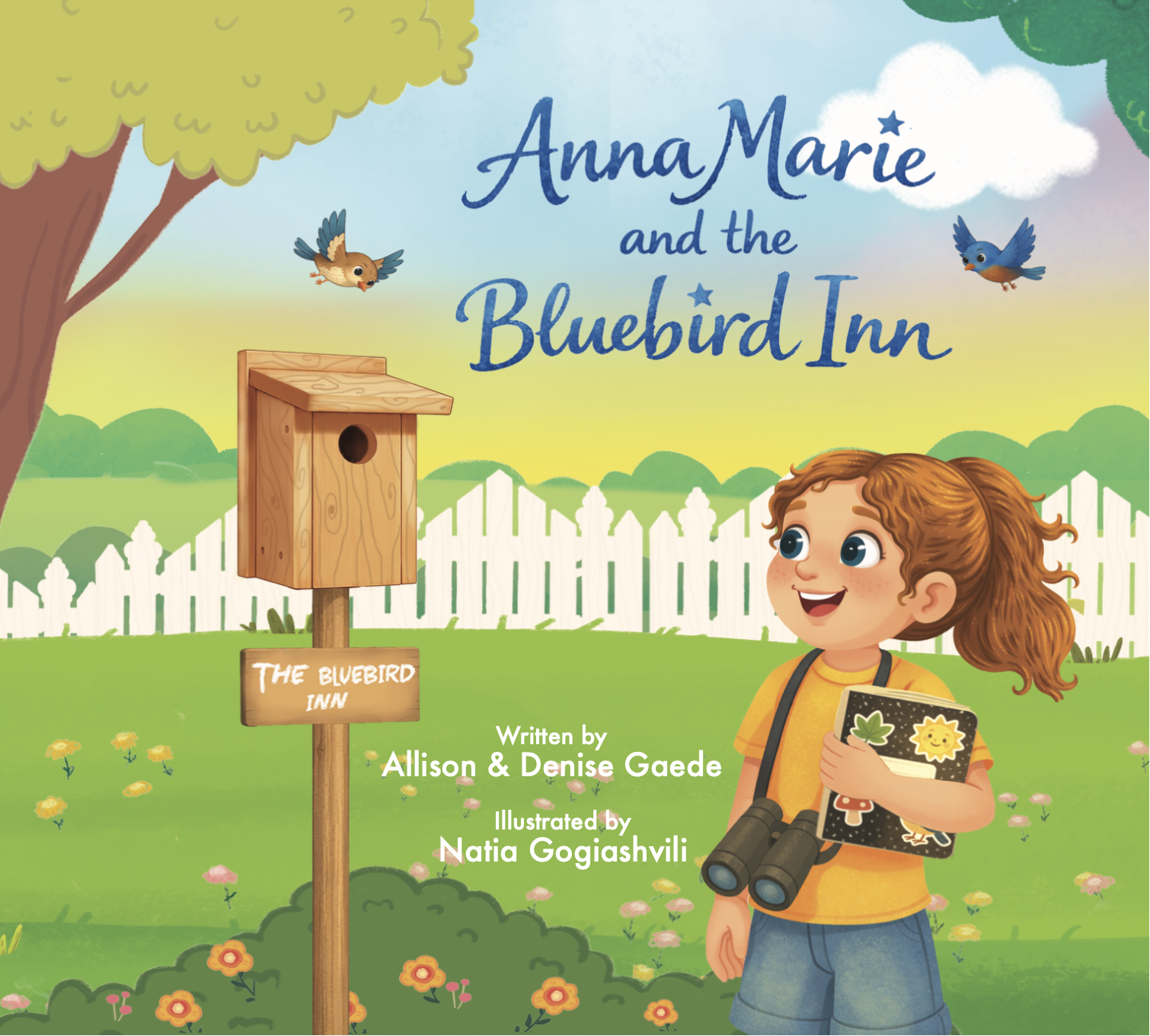 Cover of a children's book titled 'Anna Marie and the Bluebird Inn.' The scene shows a girl with red curly hair, holding a notebook, standing outdoors with a white picket fence, trees, and colorful flowers. Two bluebirds are flying in the sky, and a wooden birdhouse is mounted on a post labeled 'The Bluebird Inn.'