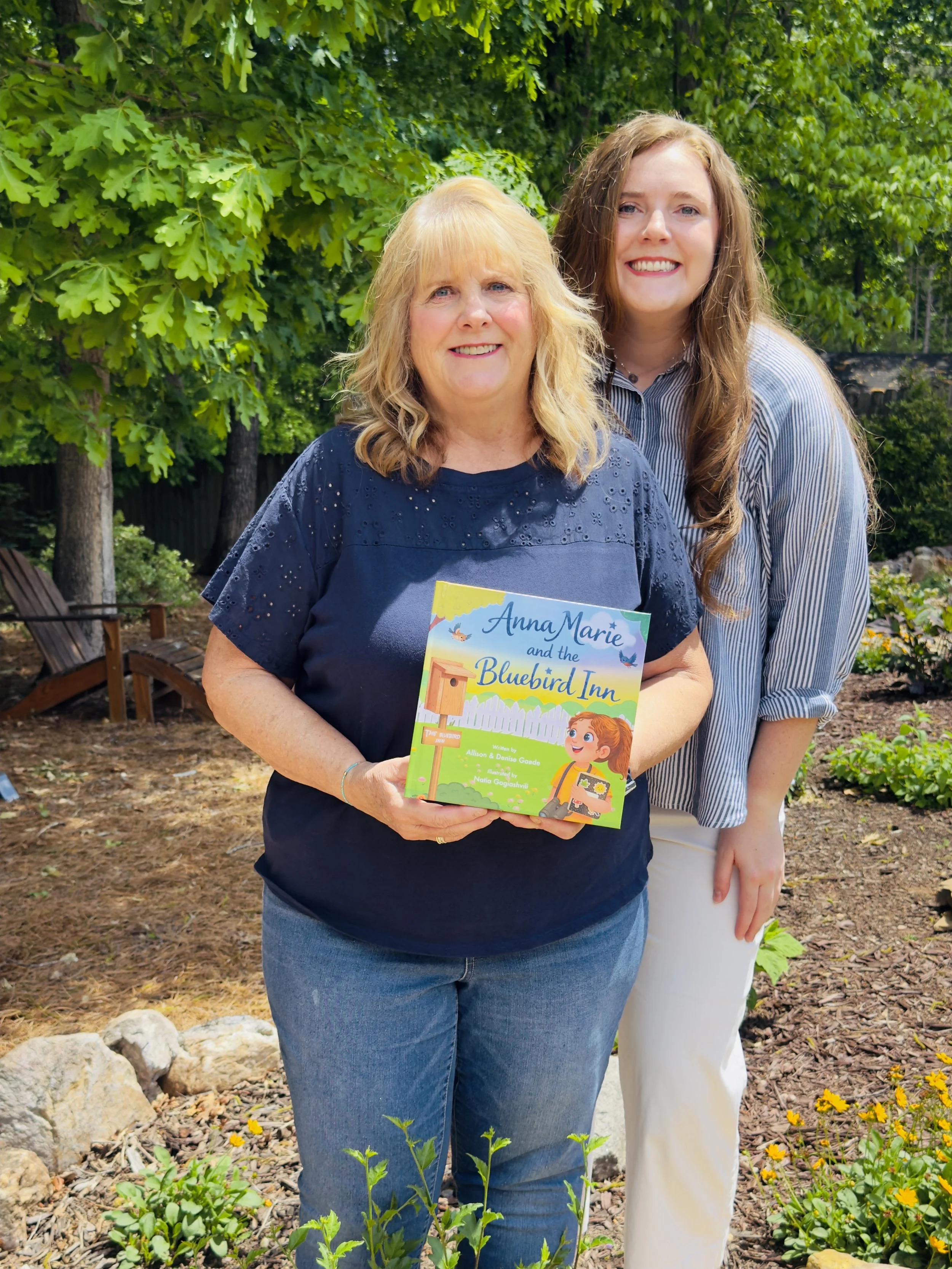 Two women standing outdoors in front of green trees, smiling, one holding a children's book titled "Anna Marie and the Bluebird Inn."