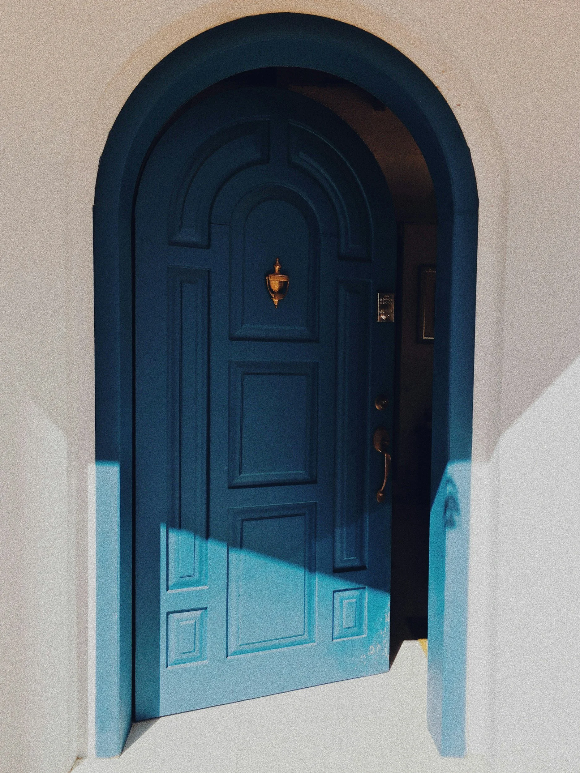 Blue arched front door with decorative panels, brass doorknob, lock, and a gold door knocker, set in a white wall.