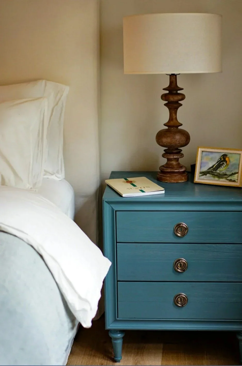 A bedroom nightstand with a blue color, three drawers with metallic knobs, a decorative wooden lamp, a white notebook with a green and red bookmark, and a small framed bird painting on top.