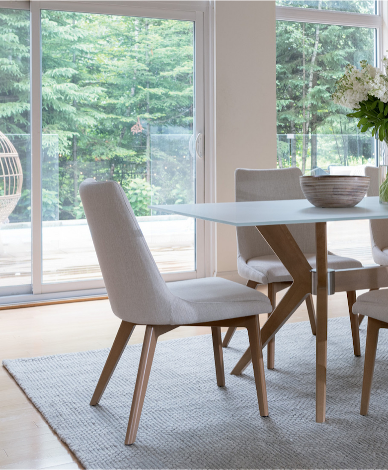 Dining area with a white table, beige upholstered chairs, a vase with white flowers, and large sliding glass doors overlooking a green wooded outdoor space.