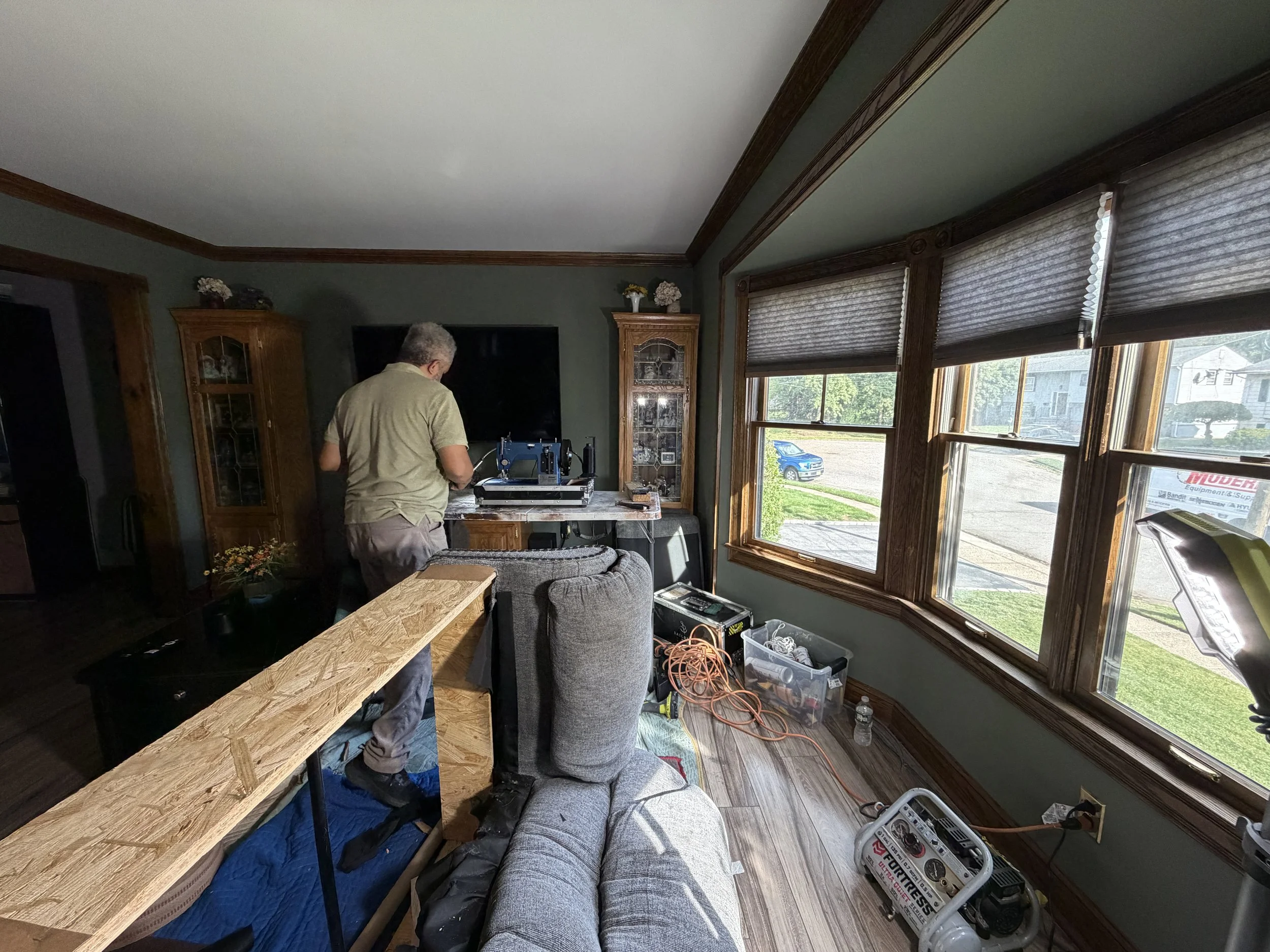 A man working on a woodworking project in a living room with large windows, a TV, and wooden furniture.