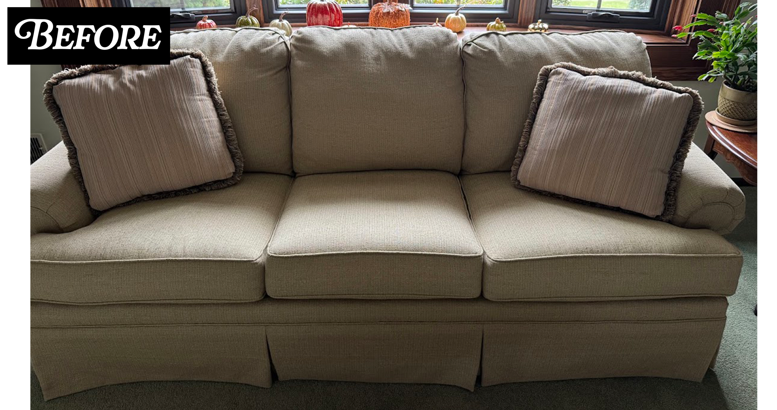 A beige upholstered sofa with three back cushions and two matching throw pillows, placed in front of a window decorated with small pumpkins on the window sill, and a potted plant on a small side table to the right.