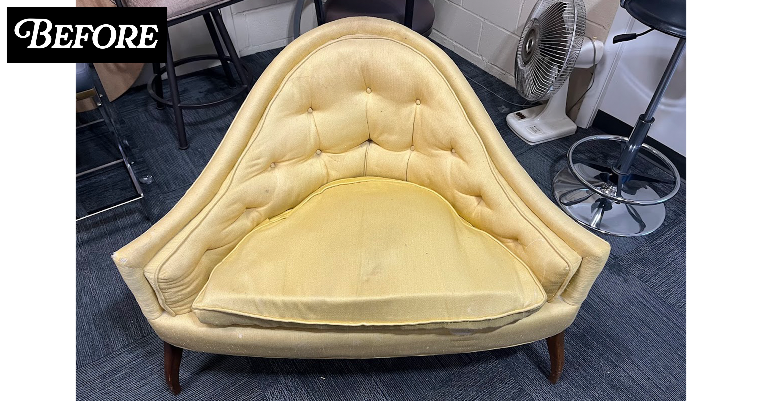A vintage yellow upholstered armchair with tufted buttons and wooden legs, situated on a dark carpeted floor in a room with various furniture and a fan.