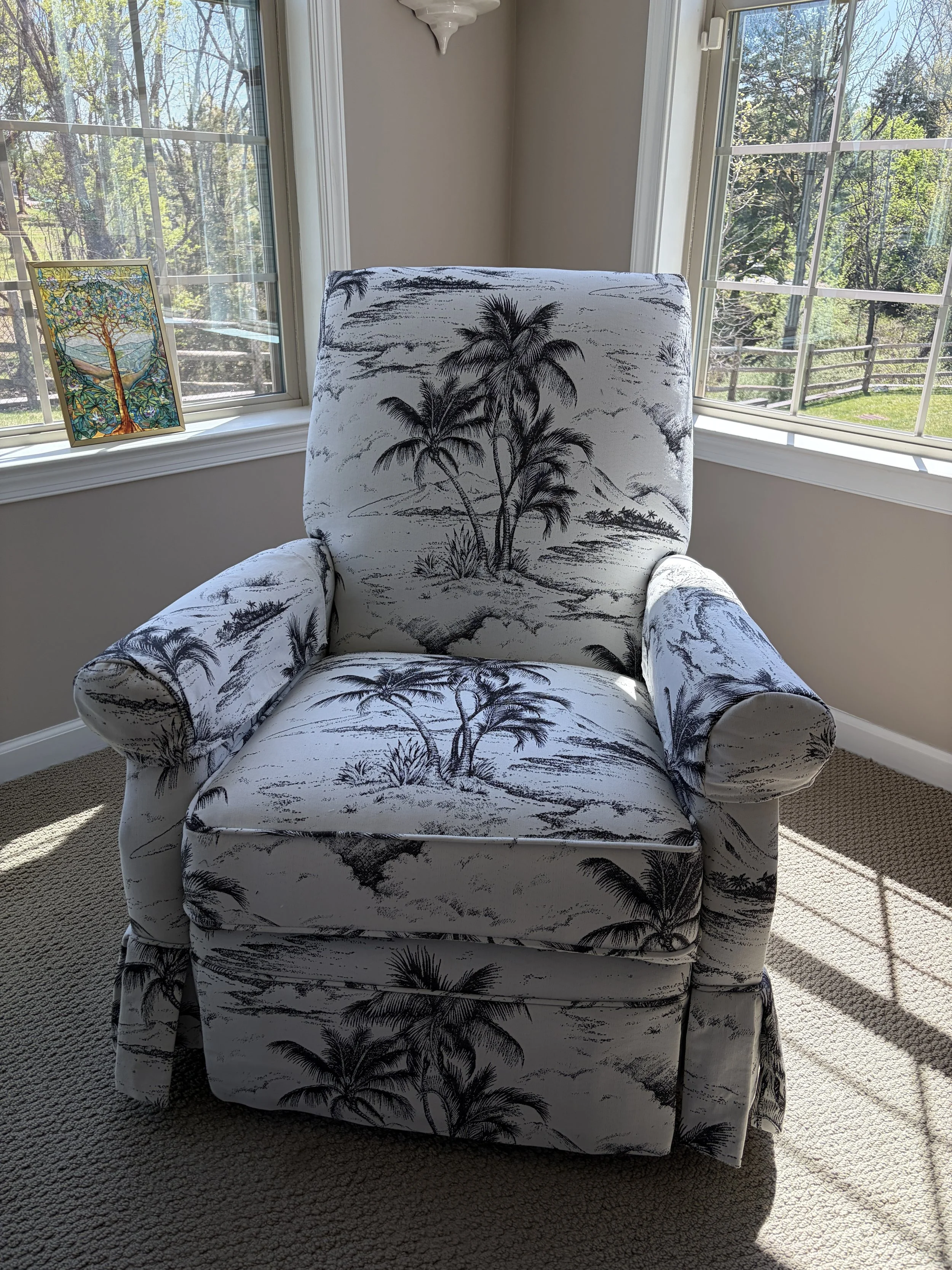 A room with a armchair featuring a black and white palm tree print, placed near a window with a stained glass artwork of a tree on the sill, sunlight streaming in through the window.