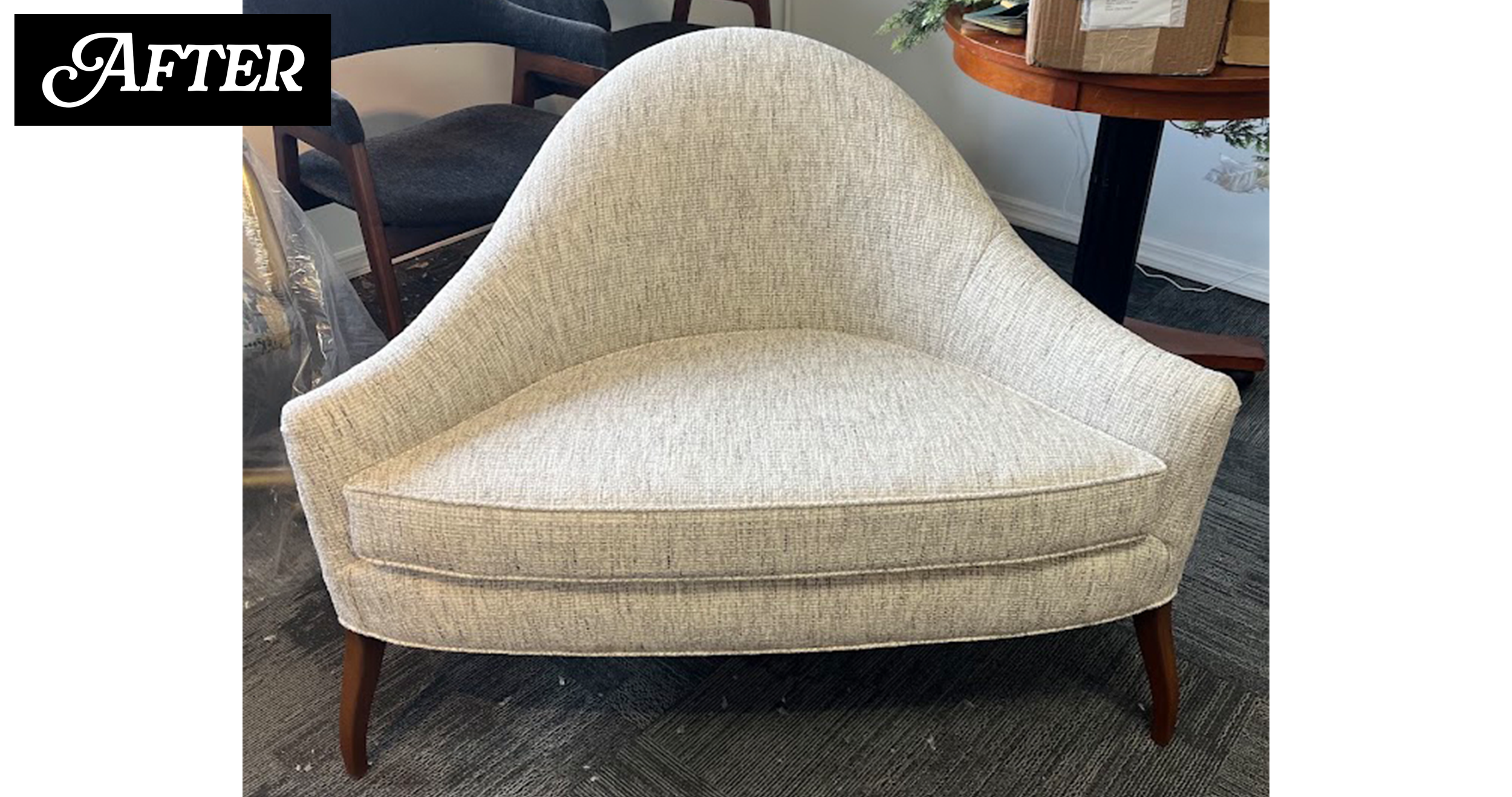 A beige upholstered armchair with wooden legs in a room, with a dark-colored chair, a round wooden table with boxes and greenery on top, and a textured dark carpet.
