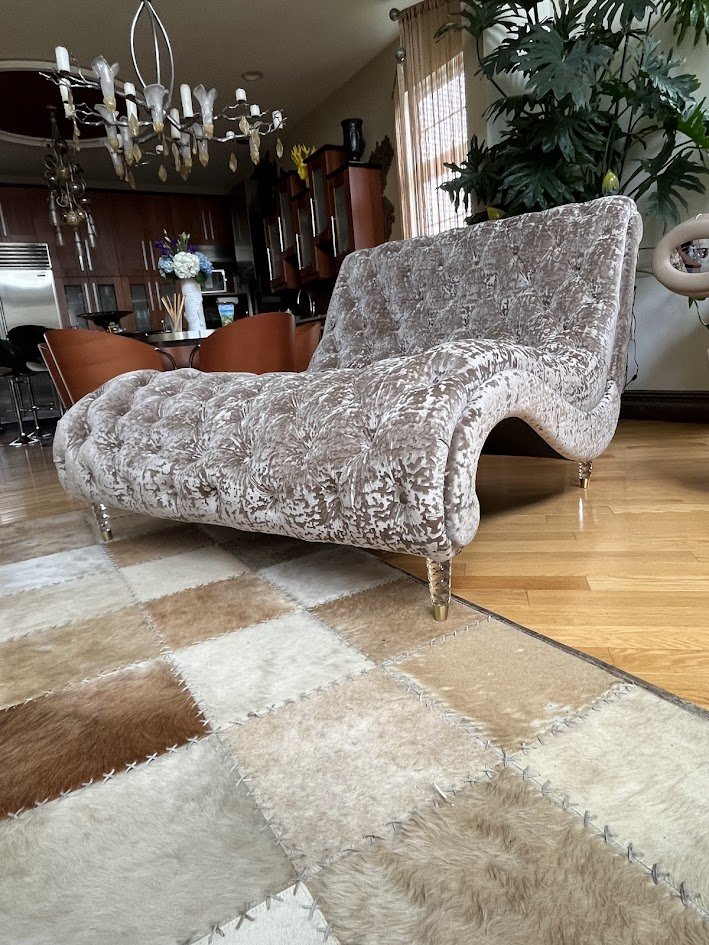 A vintage, beige, velvet tufted chaise lounge with ornate golden legs placed on a patchwork cowhide rug in a living room.