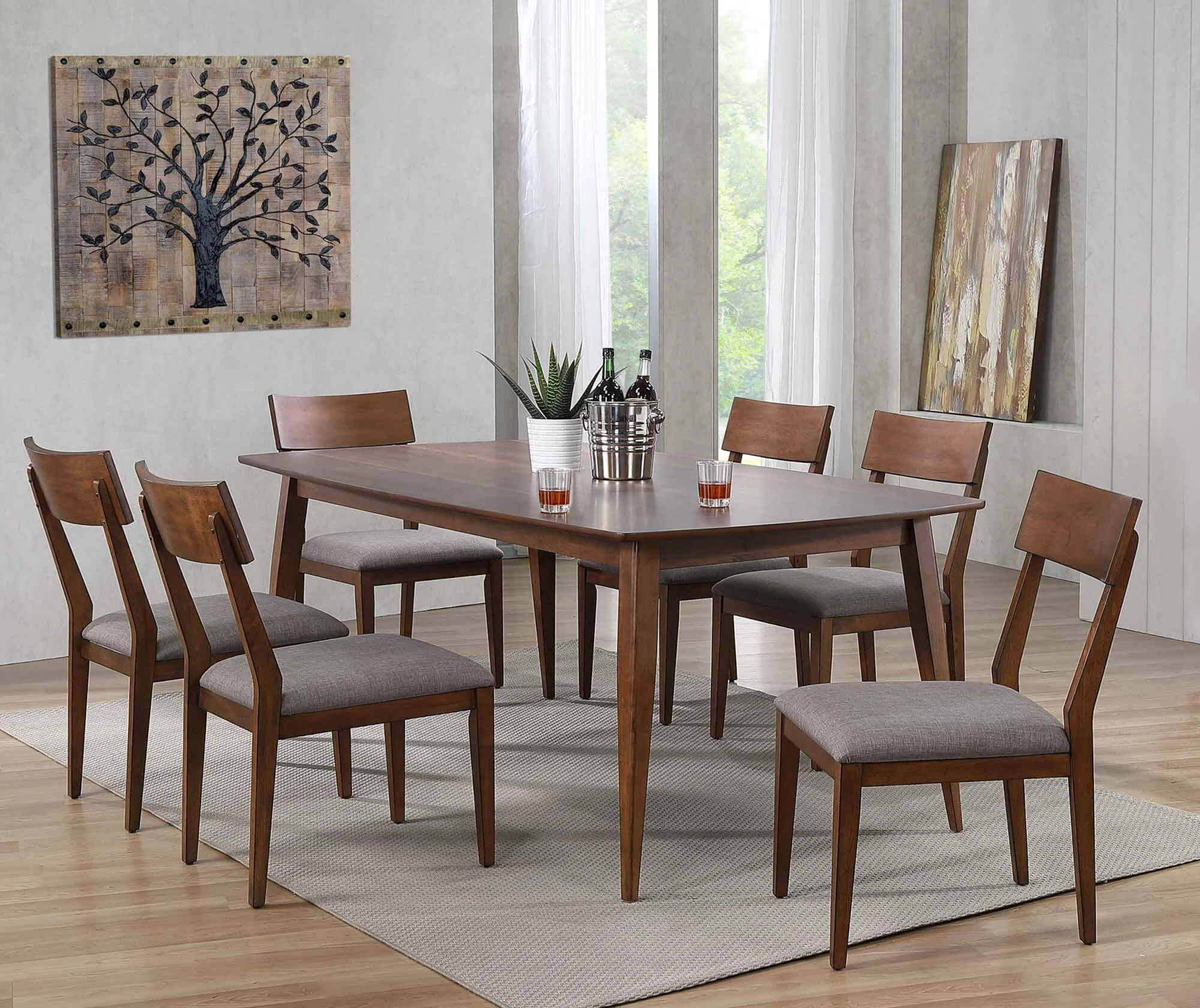 A wooden dining table with six matching chairs around it, set in a bright room with large windows, artwork on the wall, a potted plant, two bottles, three glasses, and a light rug underneath.