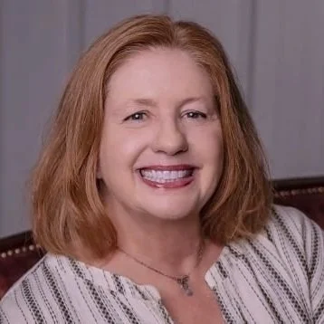 A woman with shoulder-length red hair smiling, wearing a striped blouse and a necklace, sitting indoors in front of a neutral background.