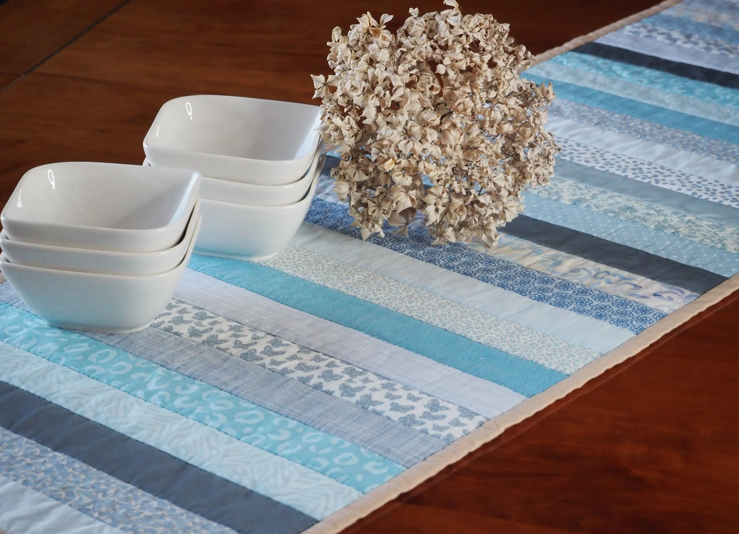 Stack of white ceramic bowls on a blue and white striped table runner with dried flowers on a wooden table.