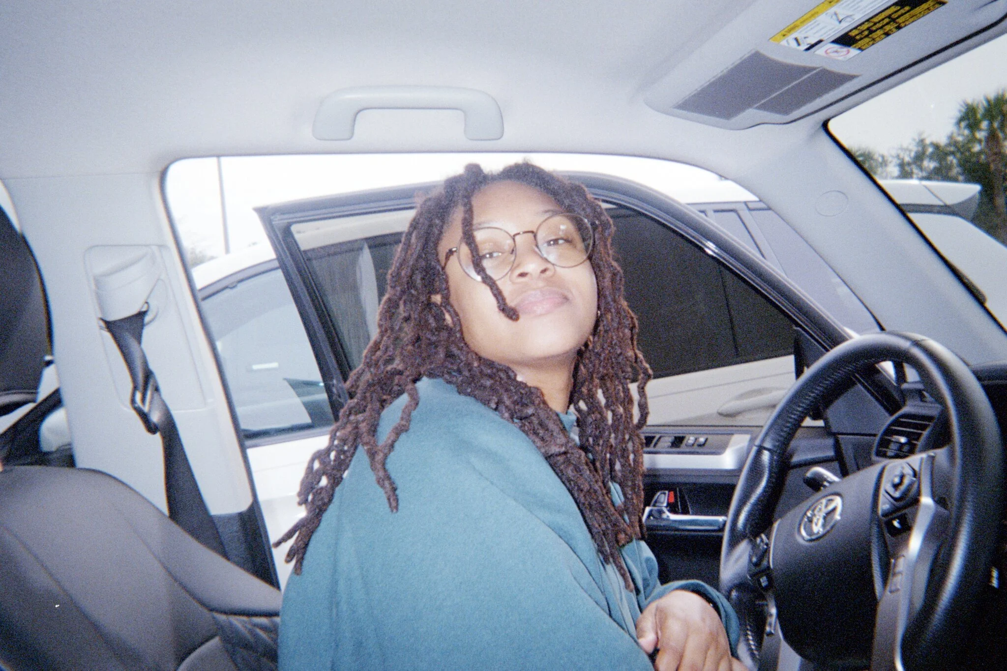 Jahnna Blyden with glasses and long dreadlocks sitting in the driver's seat of a car, looking at the camera with a slight smile.