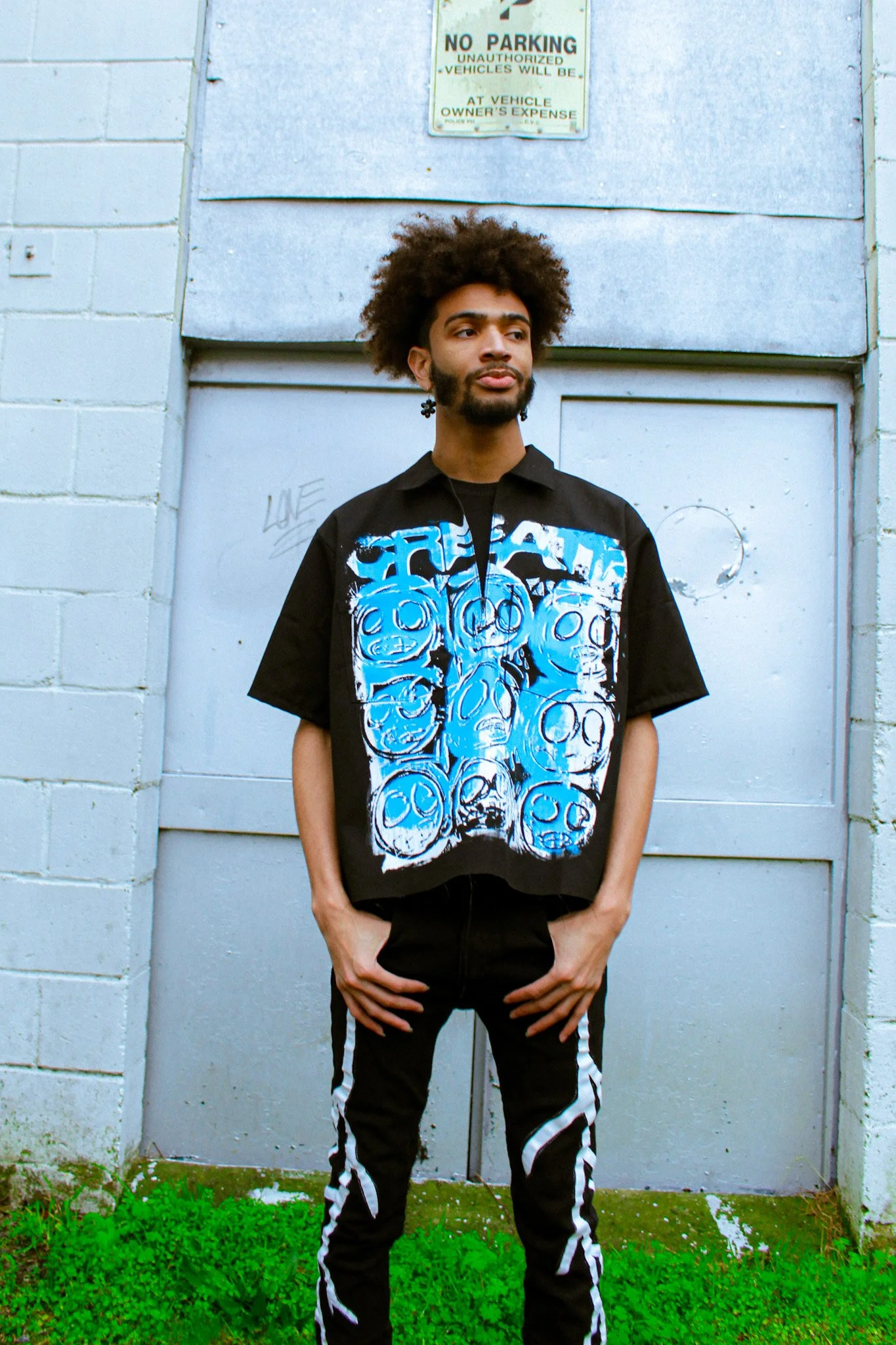 A young man with an afro hairstyle and beard standing in front of a gray metal door with graffiti, wearing a black shirt with blue graphic art and black pants with striped white details, outdoors on a grassy area.