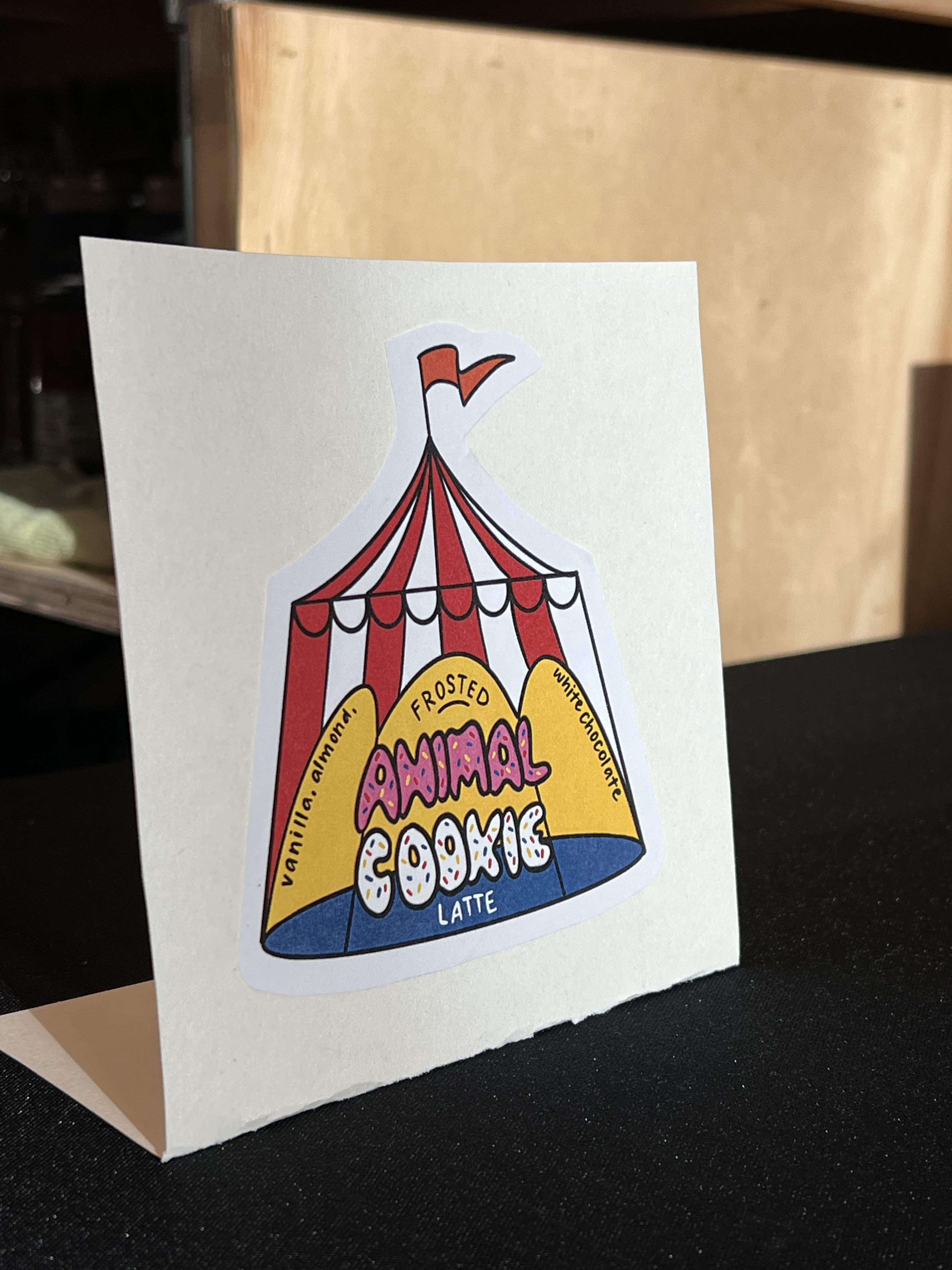 A sticker on a white card depicts a colorful circus tent with a red and white striped pattern. The tent features a yellow banner that says "Frosted Animal Cookie Latte" with flavor notes of vanilla, almond, and white chocolate.