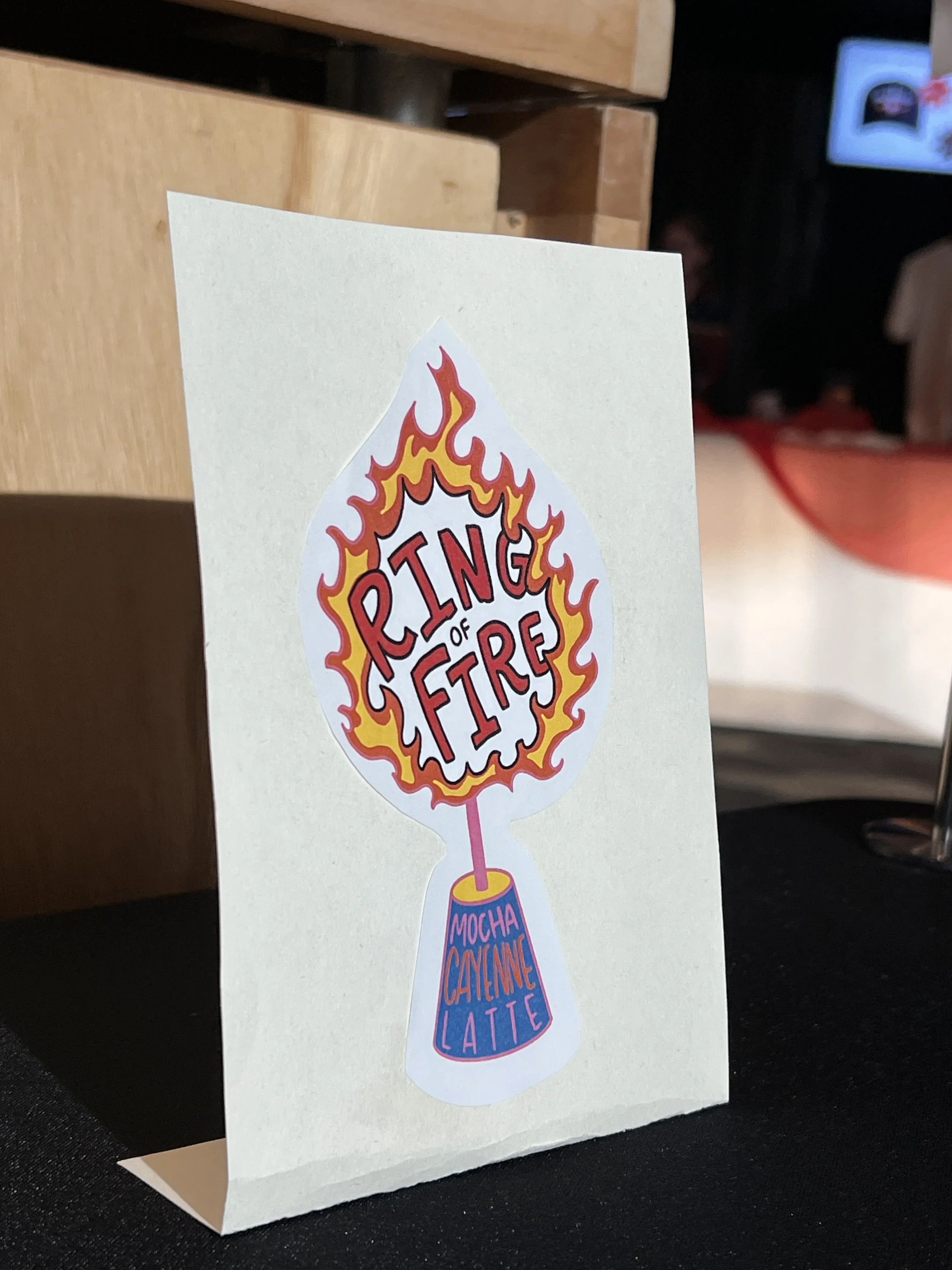 A card with a flame-shaped graphic containing the words 'Ring of Fire' surrounded by flames, and a small cup at the bottom labeled 'Mocha Cayenne Latte'.