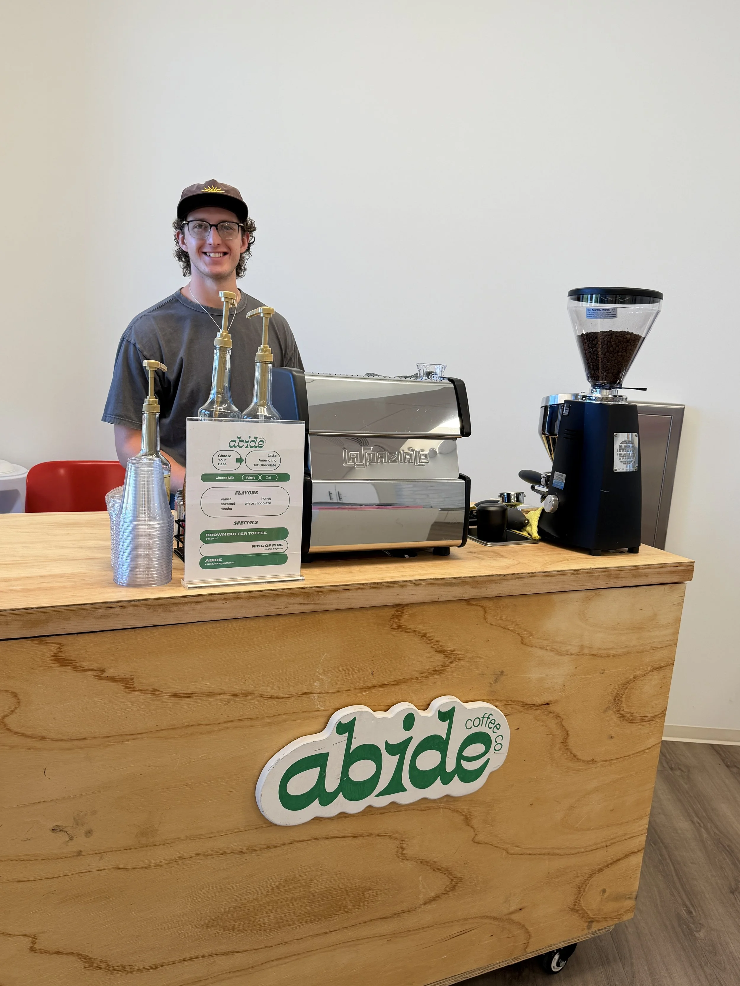 A smiling young man with glasses and a cap stands behind a wooden counter with a coffee shop sign that reads 'abide coffee co.' The counter holds bottles, a espresso machine, a coffee grinder with beans, and a menu sign.