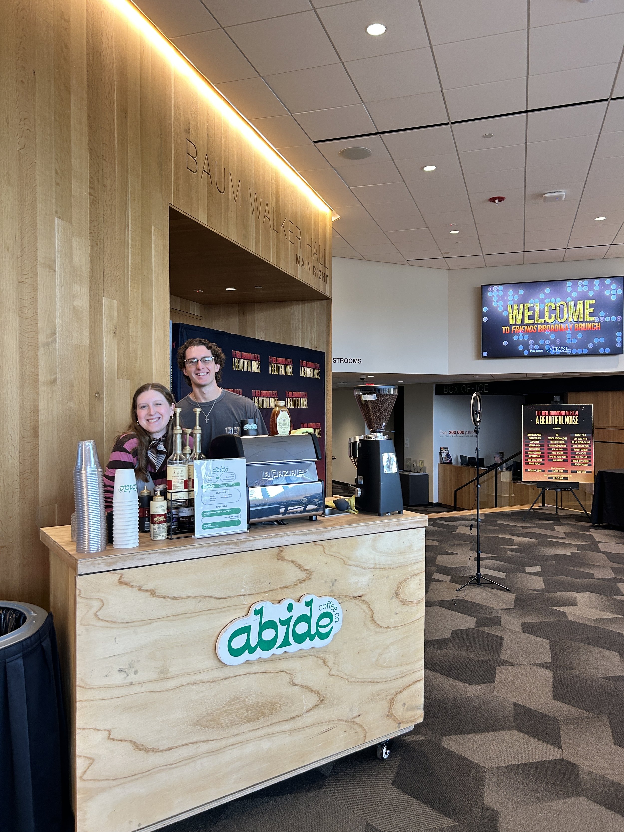 Two smiling people standing behind a coffee cart with a sign that says 'abide coffee'. The cart is in a lobby or event space with a wood-paneled wall and a large screen displaying a welcome message in the background.