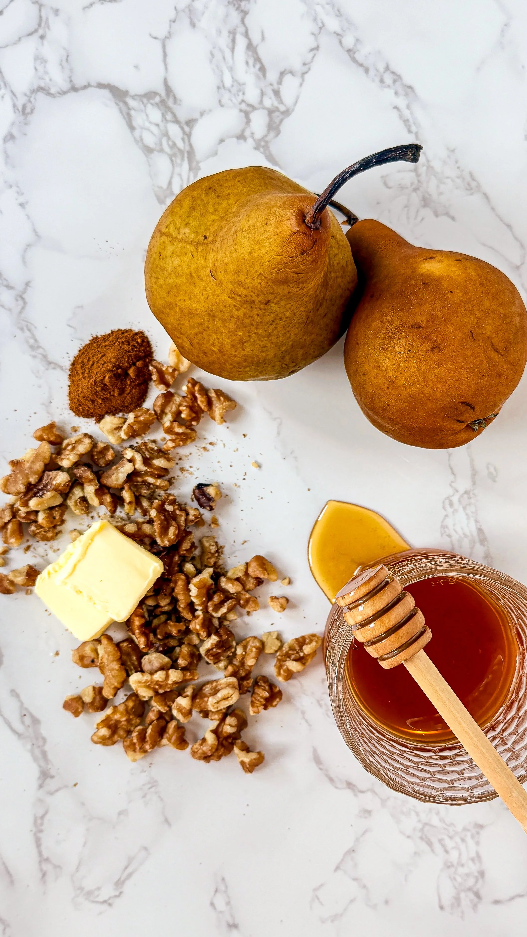 A bright, minimalist flat-lay of recipe ingredients including firm Bosc pears, a jar of raw unfiltered honey, a bottle of mild extra virgin olive oil, and a bowl of ground cinnamon on a white marble surface.