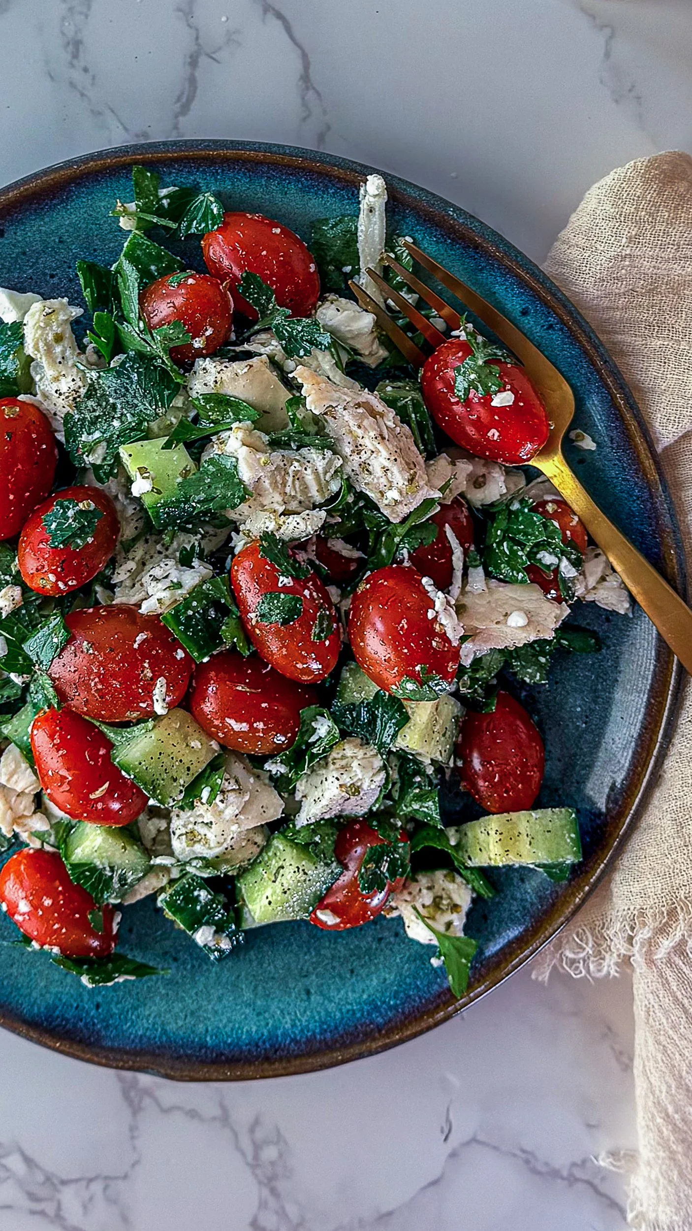 A vibrant close-up of a Mediterranean chicken salad in a dark blue bowl, featuring chunks of chicken, halved cherry tomatoes, chopped cucumbers, and fresh herbs, with a gold fork resting on the side.