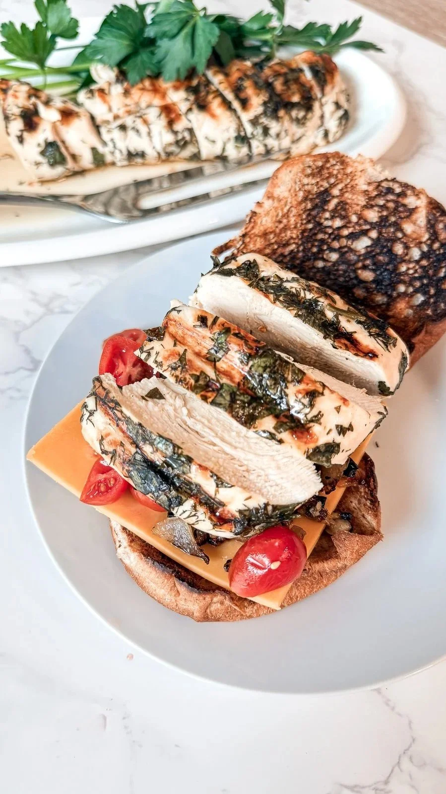 A grilled chicken sandwich on a white plate topped with herbs, tomatoes, onions, and cheddar, with a platter of sliced chicken in the background on a white marble surface.