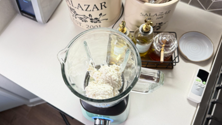 A kitchen counter with a blender of creamy mixture surrounded by utensils and ingredients, showing the preparation setup.