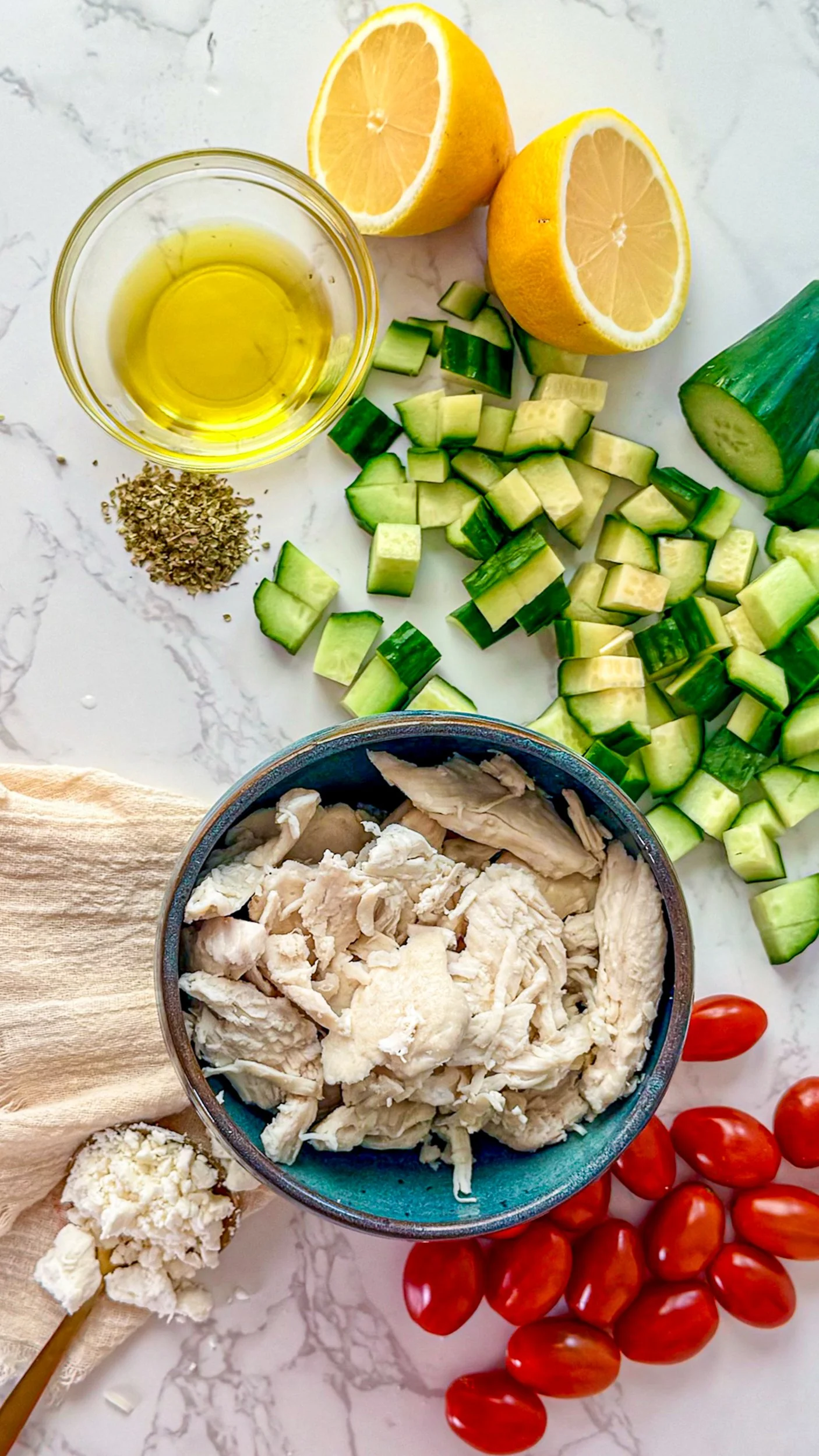 A flat lay of recipe ingredients including an english cucumber, lemon, olive oil, chicken, oregano and cherry tomatoes on a marble surface