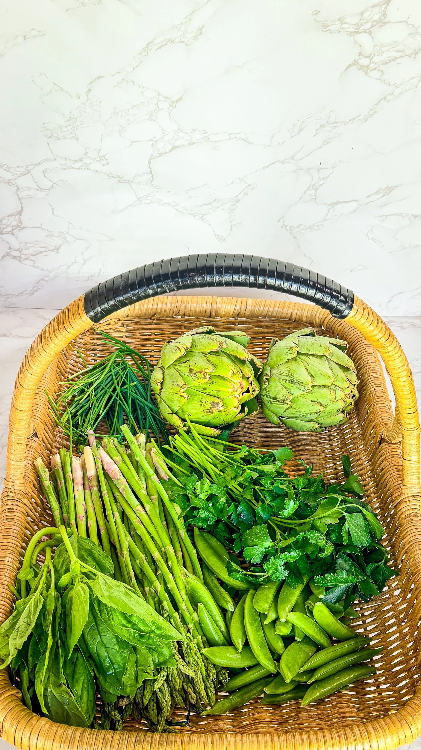 A top-down photograph of a light-colored woven wicker basket with a black-wrapped handle, resting on a white marble surface. The basket is filled with a variety of vibrant green produce,