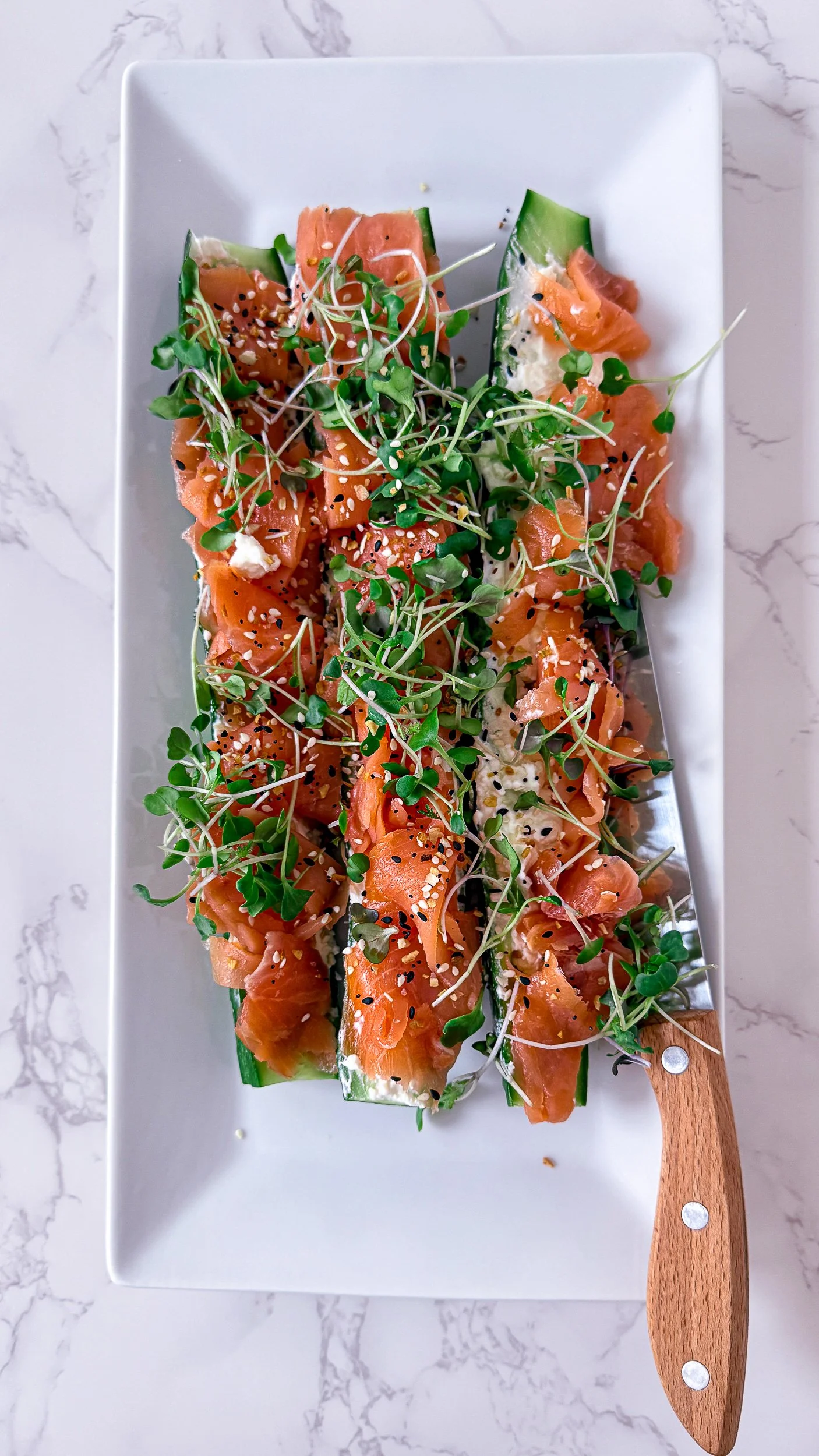 Four English cucumber boats filled with creamy whipped feta, topped with generous portions of smoked salmon and fresh microgreens on a white serving platter.
