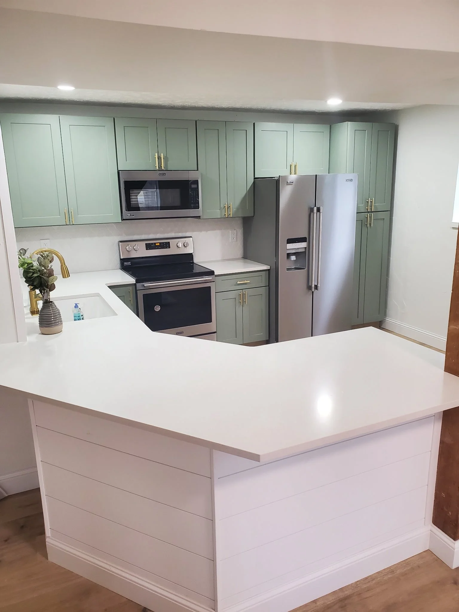 Modern kitchen with light green cabinets, stainless steel refrigerator, microwave, oven, and white countertops, with a kitchen island in the foreground.
