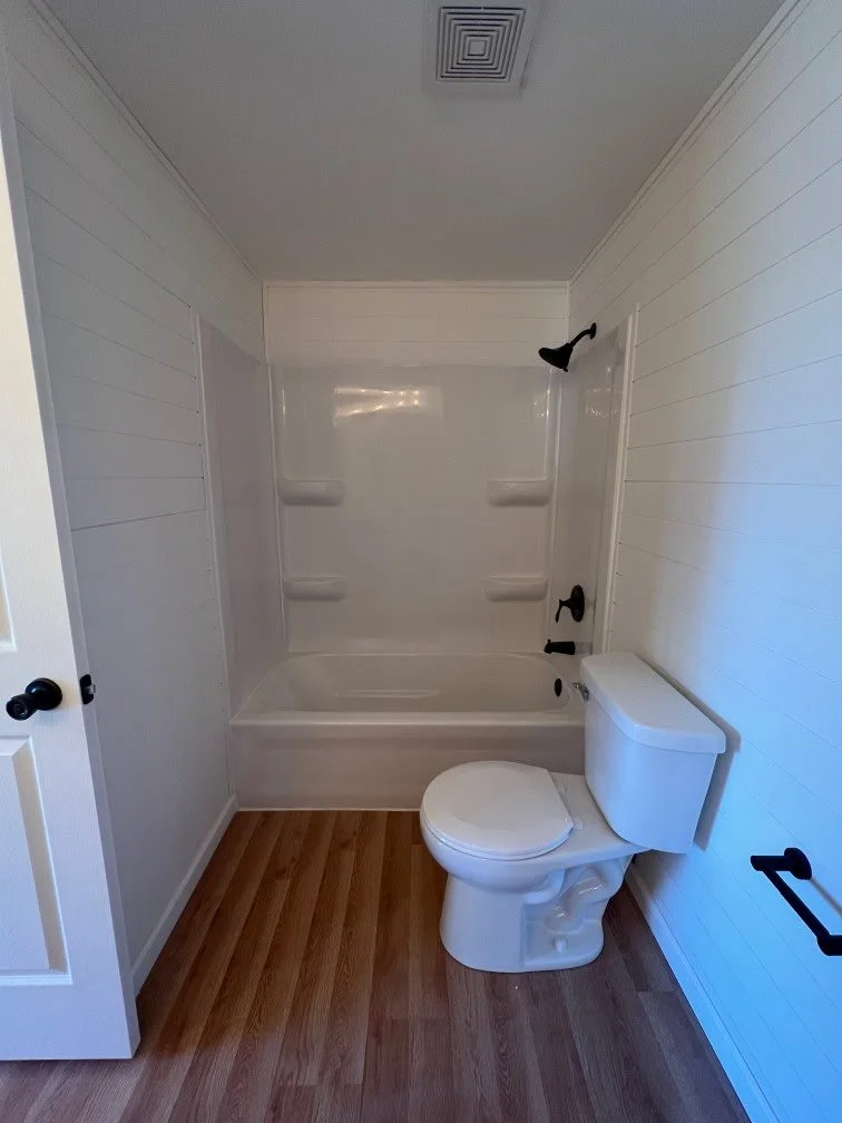 A small bathroom with a white toilet next to a bathtub with a molded shower surround and black fixtures. The bathroom has wood-like flooring and white paneled walls.