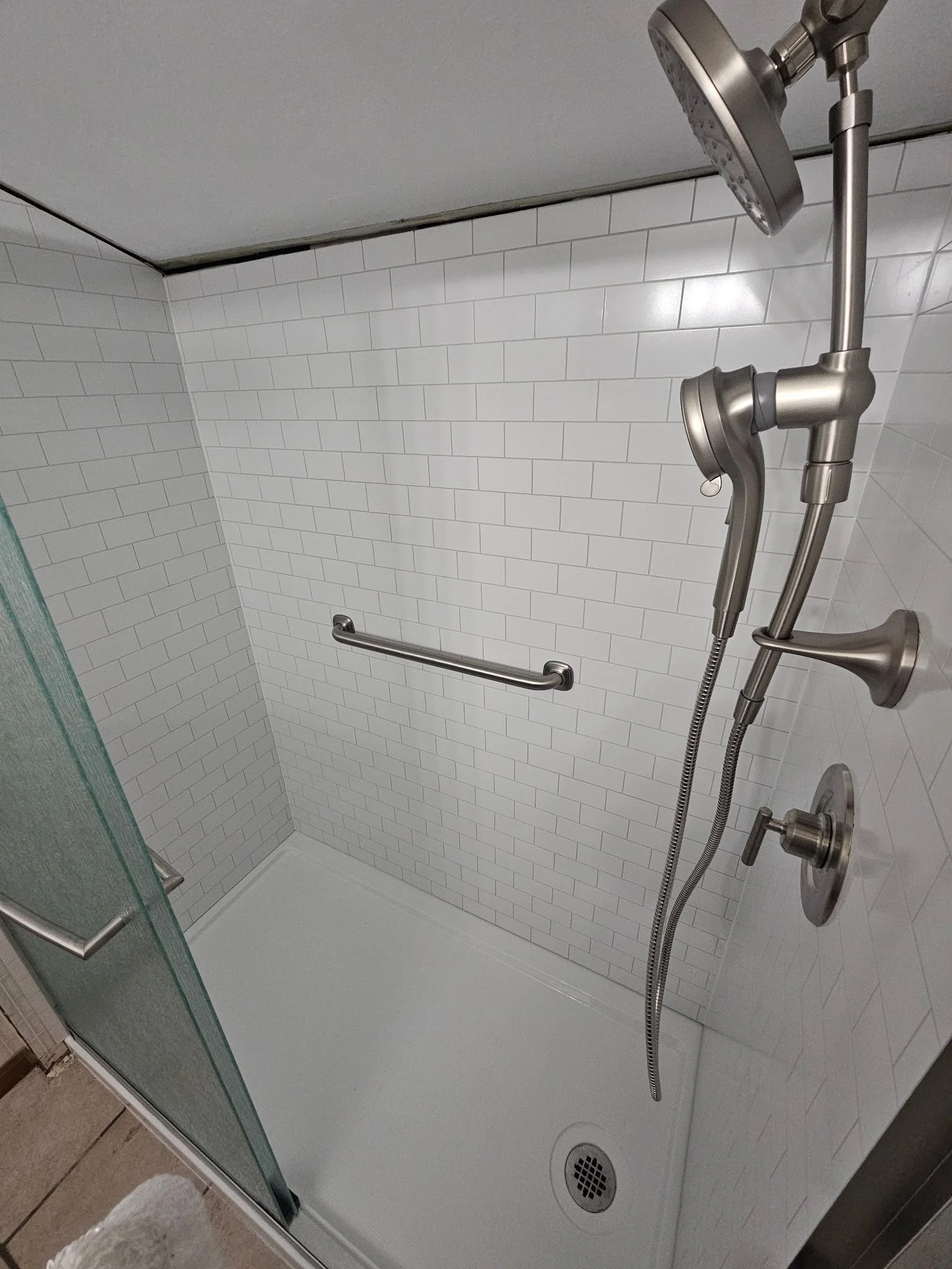 A walk-in shower with white subway tile walls, a handheld showerhead, a fixed showerhead, a grab bar, and a glass door with a towel bar, in a bathroom.