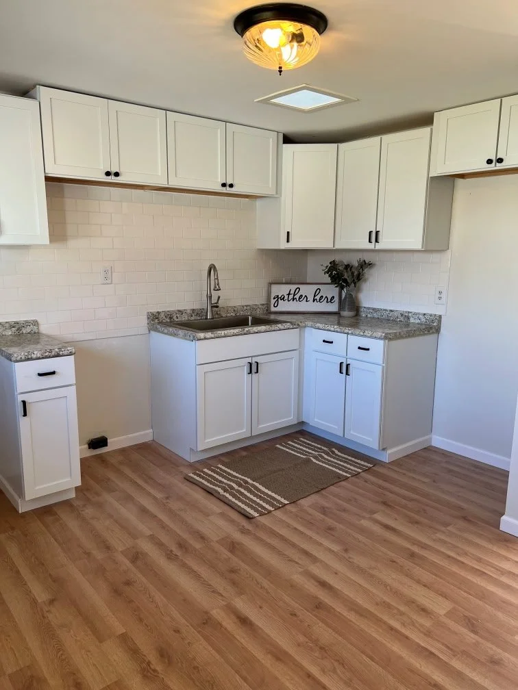 Empty kitchen with white cabinets, granite countertops, a small rug, and a decorative sign that says 'gather here' on the countertop, with a vase of flowers and a ceiling light fixture.