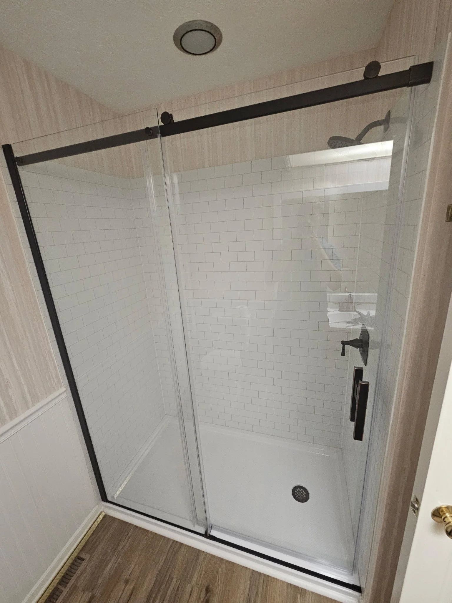 A walk-in shower with a black-framed glass door, white brick-patterned tiles on the walls, and a black showerhead and handle. The shower floor is white with a round drain.
