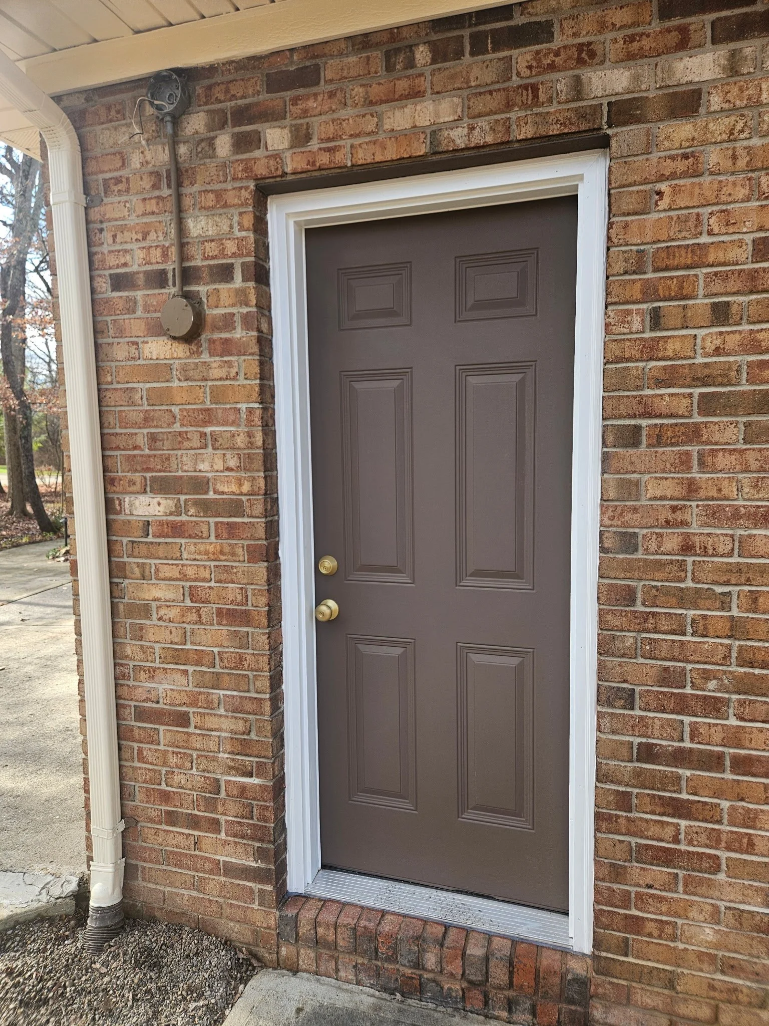 A brown front door set in a brick wall with decorative molding and a white frame, two brass doorknobs, and a small step at the base.