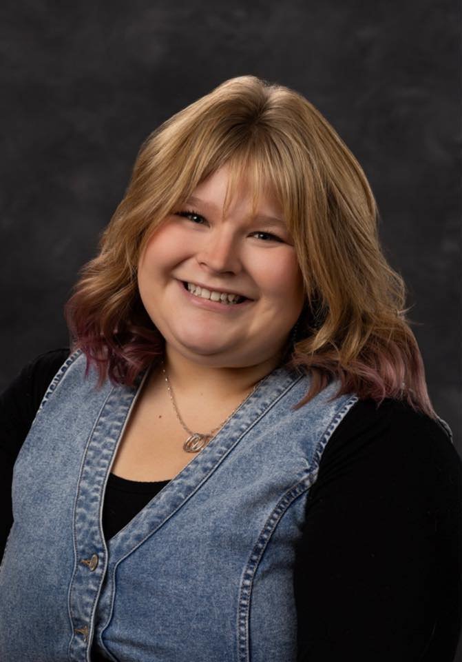 A smiling woman with shoulder-length blonde hair with purple tips, wearing a black shirt, a denim vest, and a necklace, against a dark backdrop.