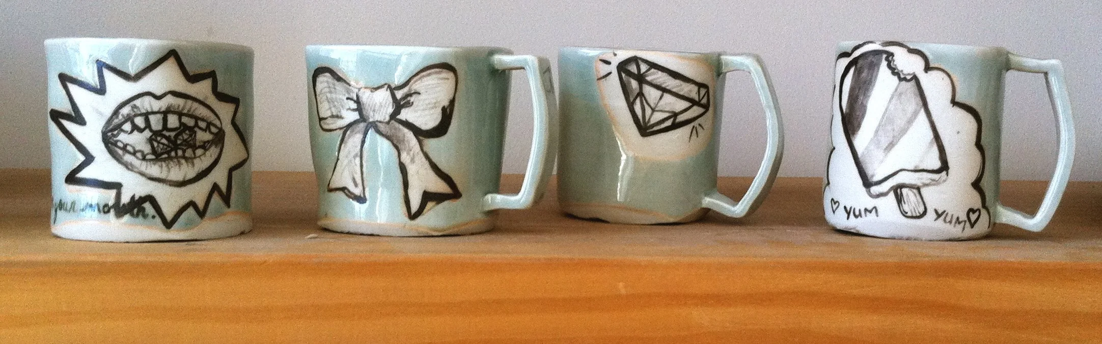 Four ceramic mugs with hand-drawn black and white illustrations on a wooden shelf. The first mug has an open mouth with a gemstone inside, the second has a bow, the third has a diamond, and the fourth has a popsicle with the words 'yum yum' and small