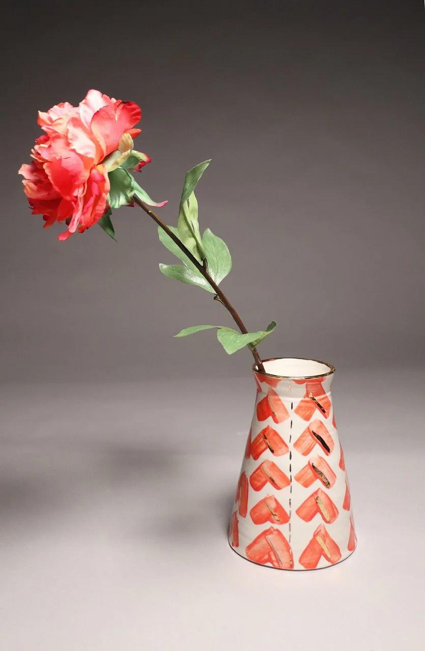 A single red and pink flower in a white ceramic vase with red heart and arrow patterns, against a plain gray background.