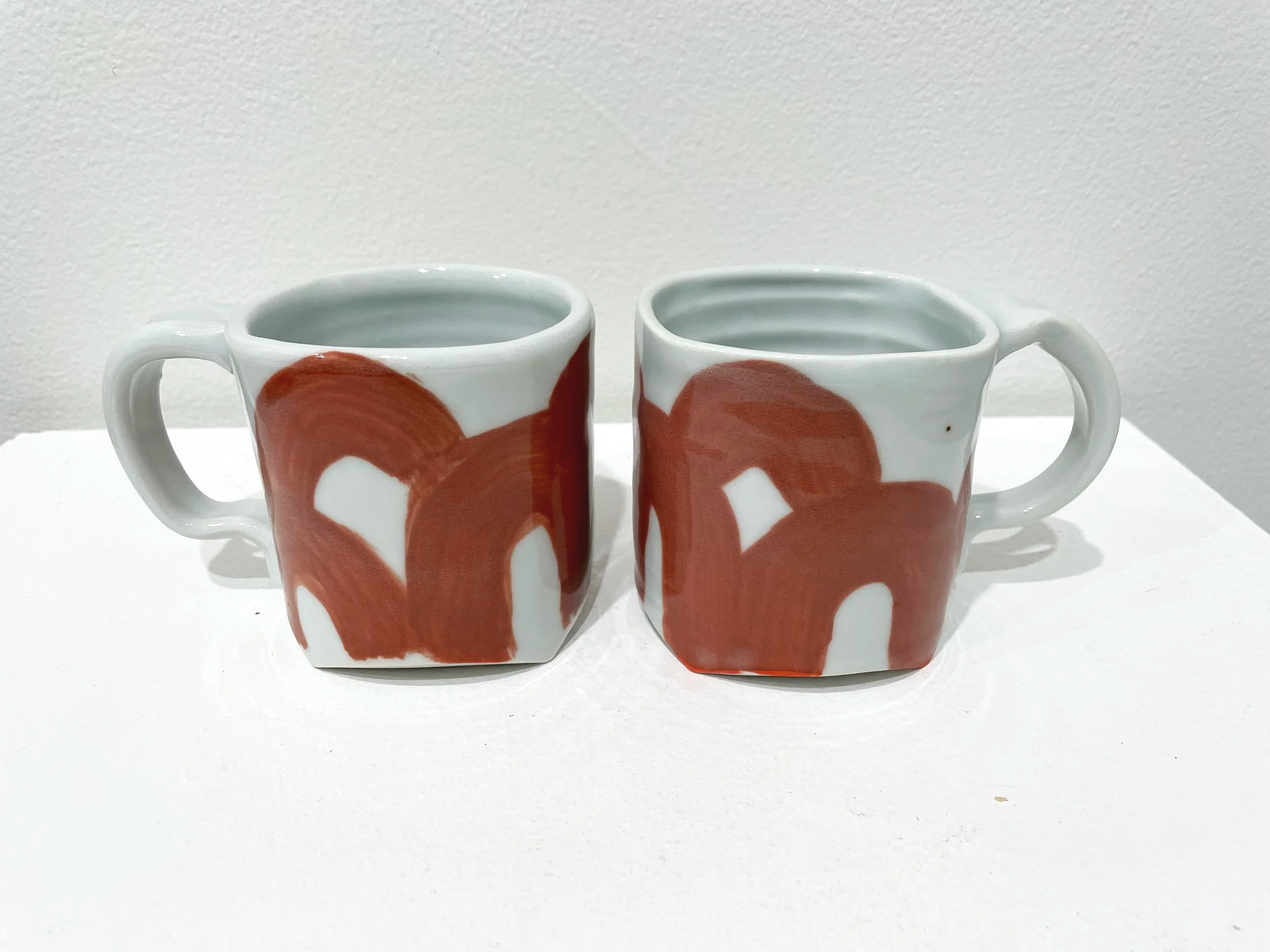 Two white ceramic mugs with abstract brown brushstroke designs, placed side by side on a white surface against a plain white wall.