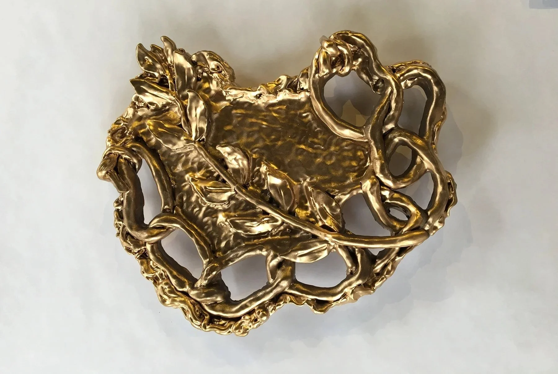 Gold-colored decorative brooch in the shape of a heart with leaves and intricate border design.