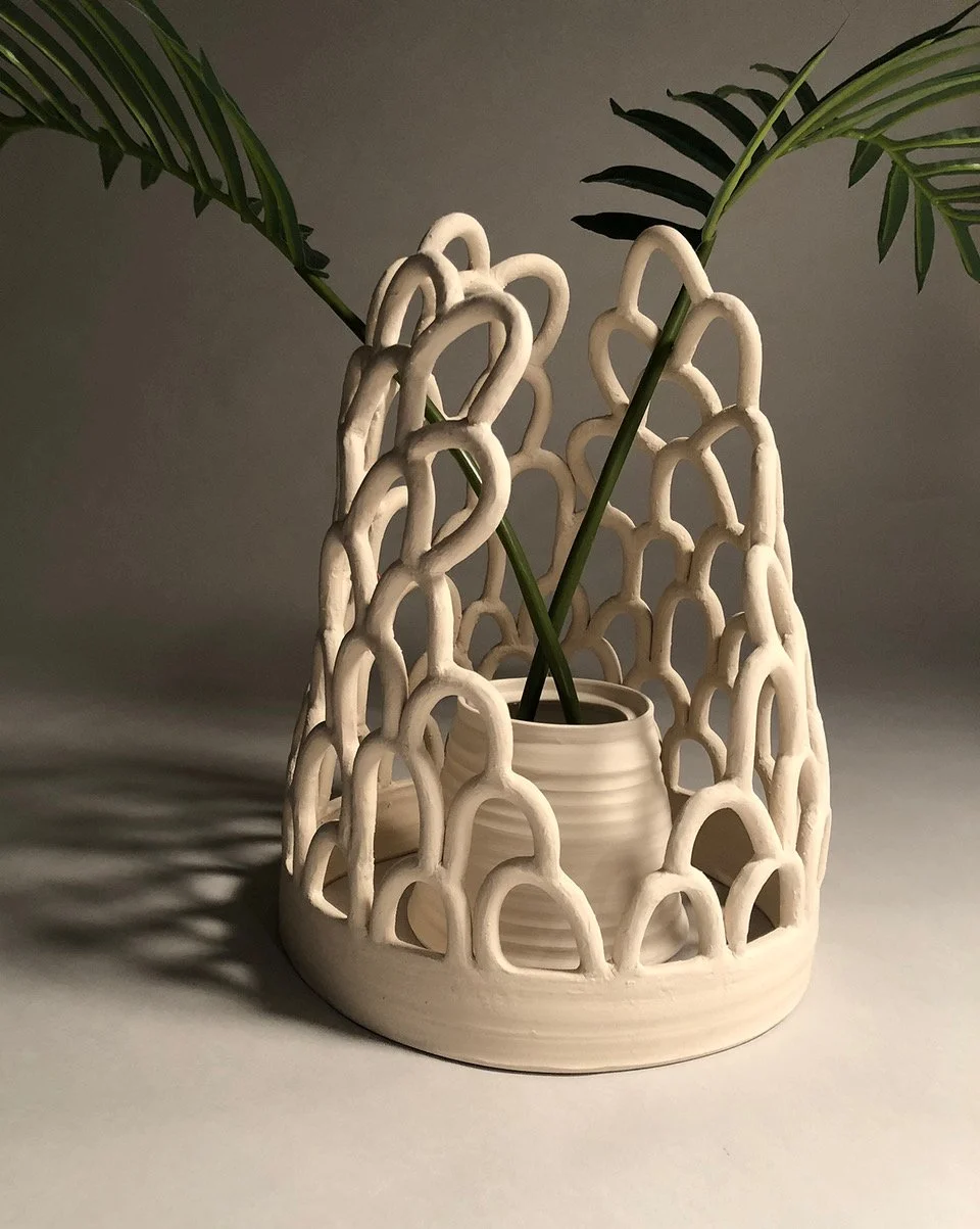 A white decorative vase with a lattice design, holding green plant stems with long, slender leaves, placed on a plain surface with soft lighting.