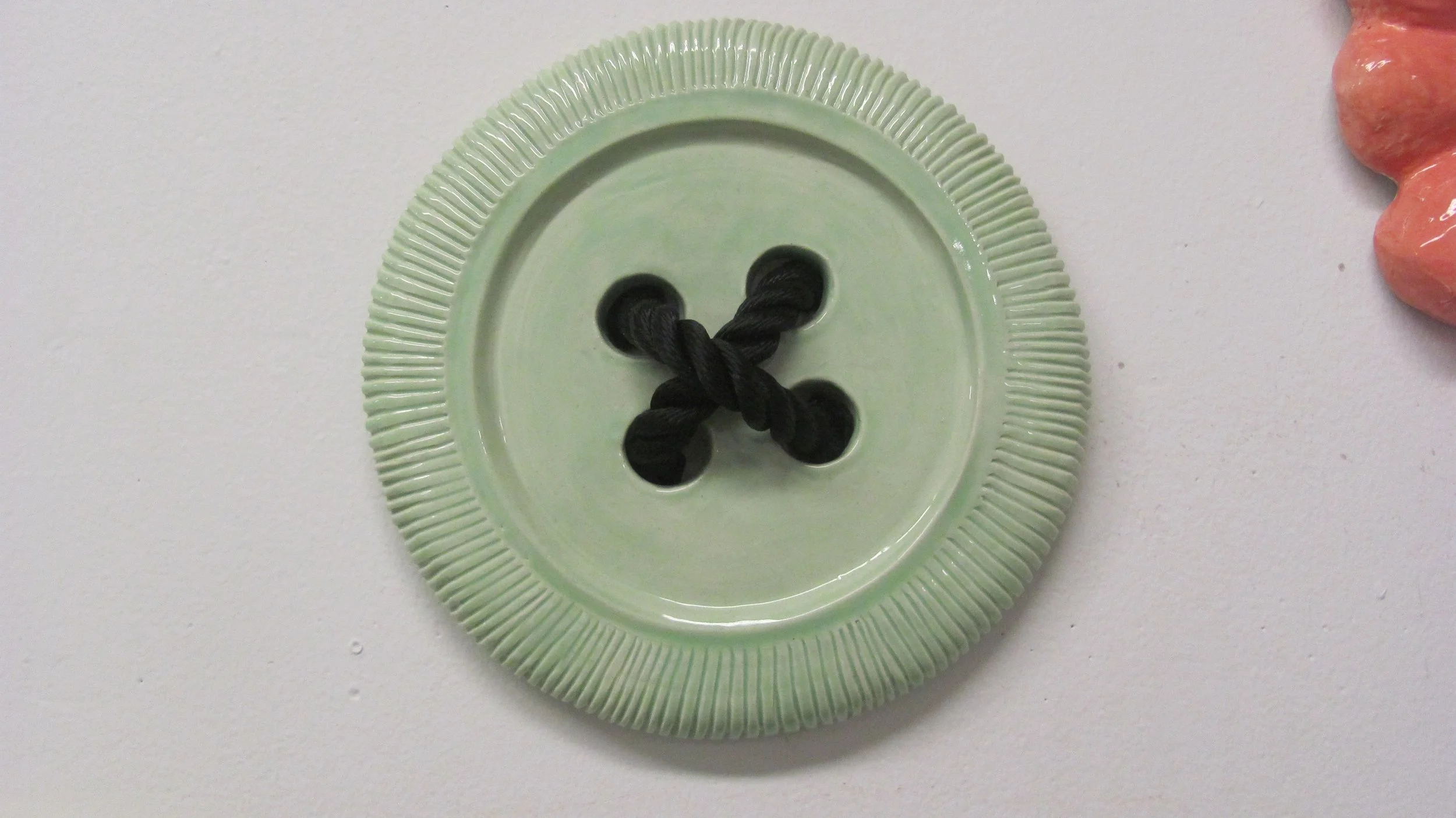 A light green ceramic button with four holes and a black twisted cord inserted through two pairs of holes, on a white surface. Part of a pink object is visible on the right edge of the image.