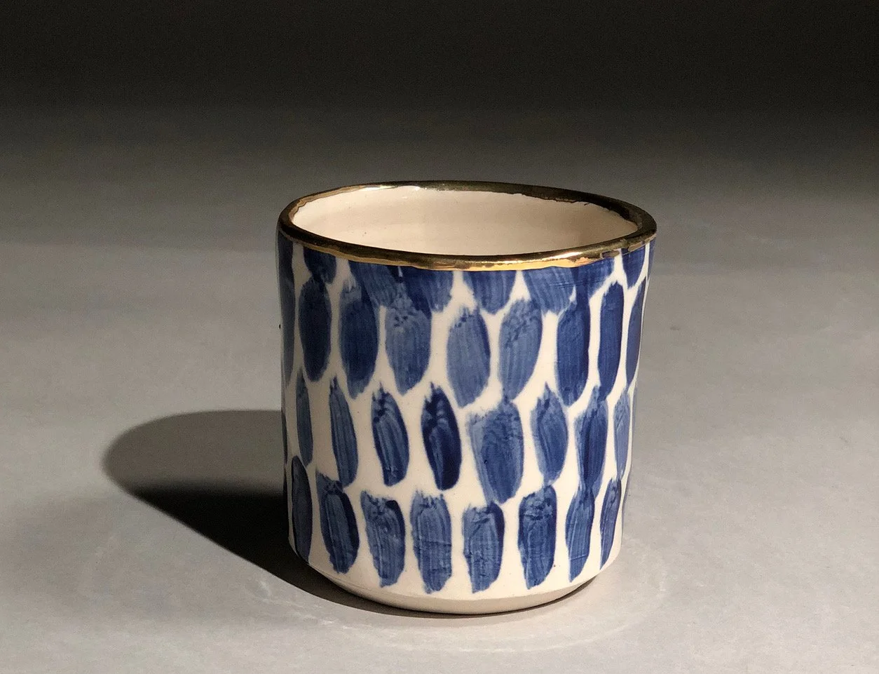 A ceramic cup with a blue and white geometric pattern and a gold rim, casting a shadow on a gray surface.