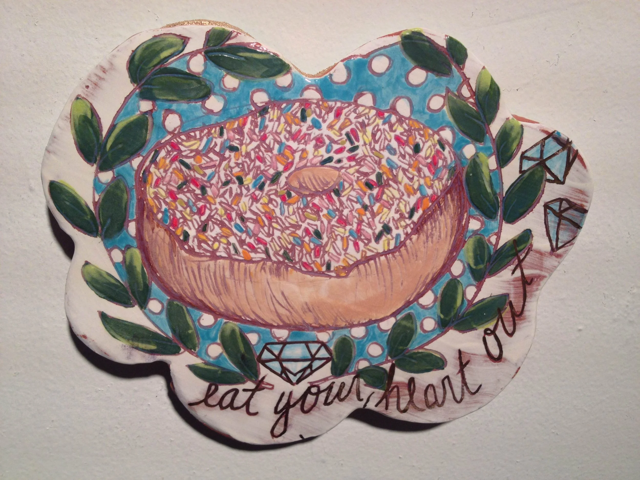 Colorful hand-painted illustration of a donut topped with rainbow sprinkles, surrounded by green leaves and two diamond illustrations, with the phrase "eat your heart" written at the bottom.