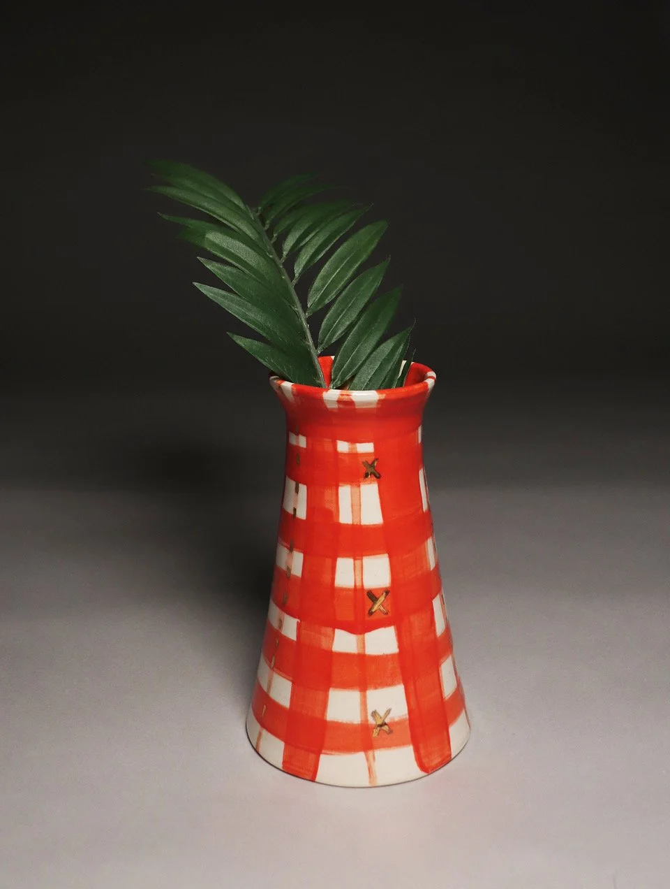 A ceramic vase with a red and white checkered pattern holding three green palm leaves against a dark background.