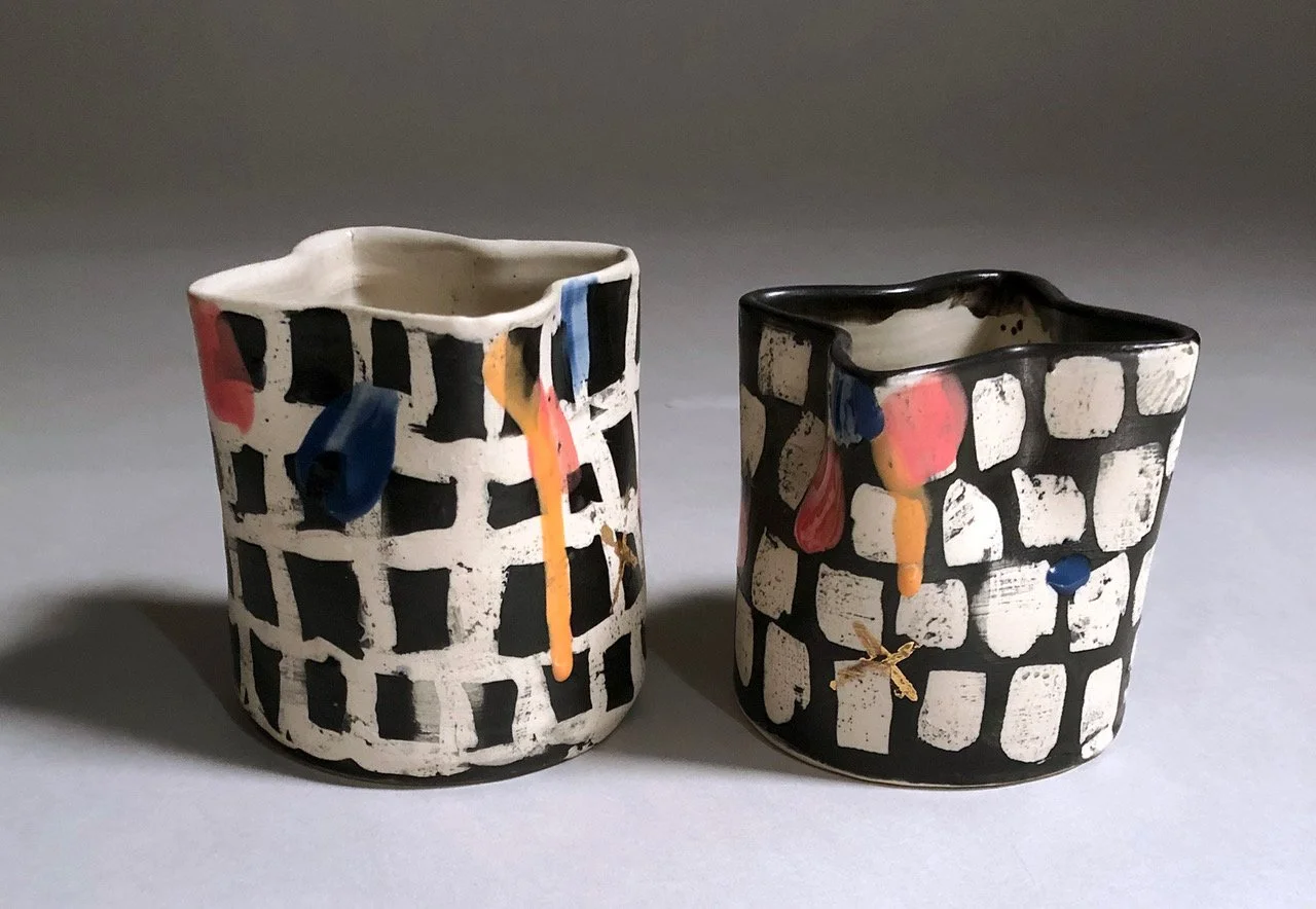 Two handcrafted ceramic cups with geometric black and white patterns, adorned with colorful abstract paint splashes in red, blue, and orange.