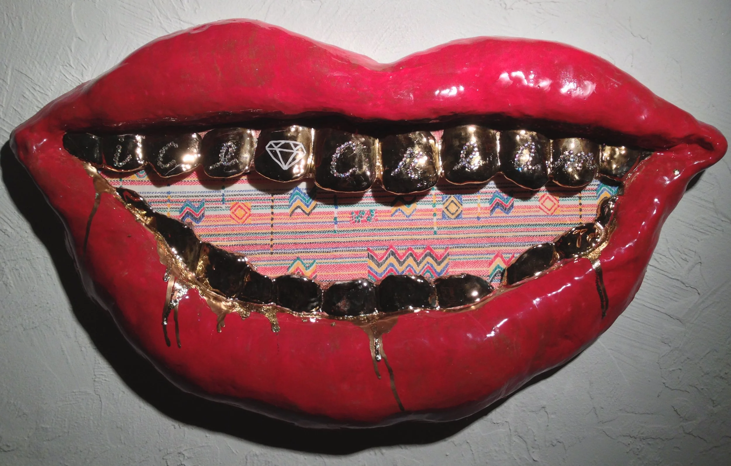 A decorative art piece of a red lips-shaped sculpture with open mouth, featuring gold and black teeth. Some teeth have embellishments, including a diamond symbol and the words 'Love' and 'Peace'. The interior of the mouth has colorful woven fabric wi