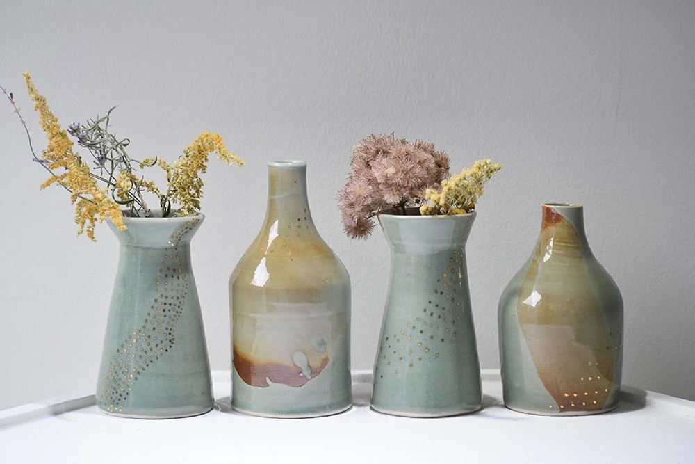 Four ceramic vases with dried flowers on a white surface against a light gray wall.