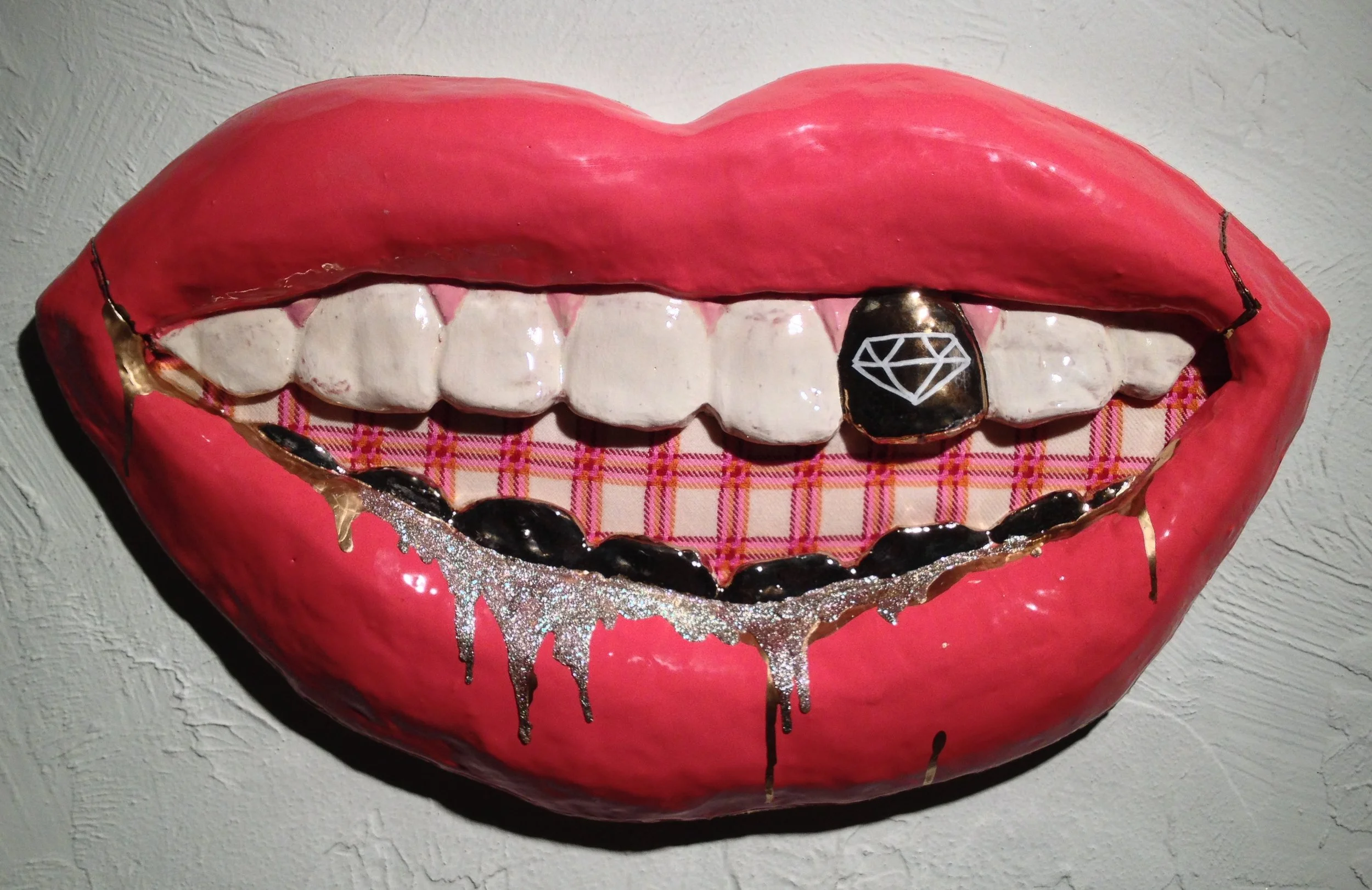 Artistic sculpture of a smiling mouth with pink lips, white teeth, and decorative elements including a black tooth with a diamond symbol, gold accents, black drips, and a plaid fabric background inside the mouth.