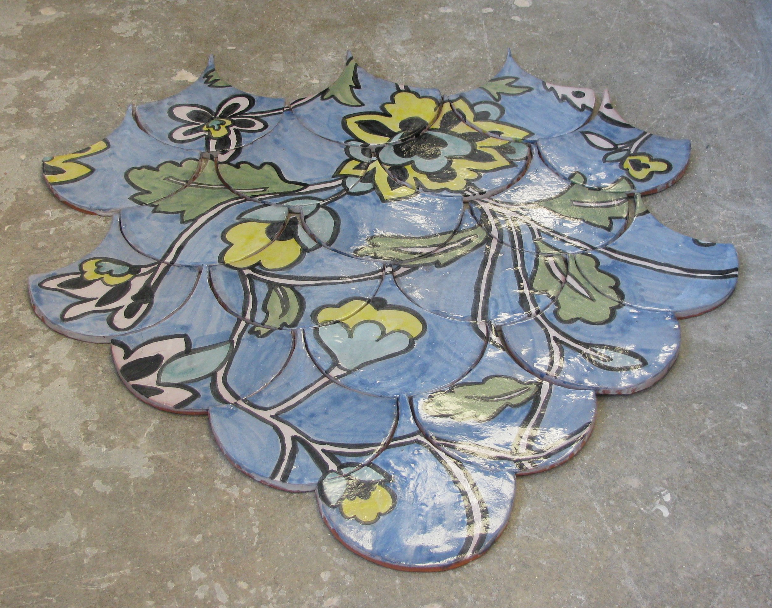 Colorful ceramic mosaic of flowers and leaves arranged in an overlapping pattern on a concrete floor.